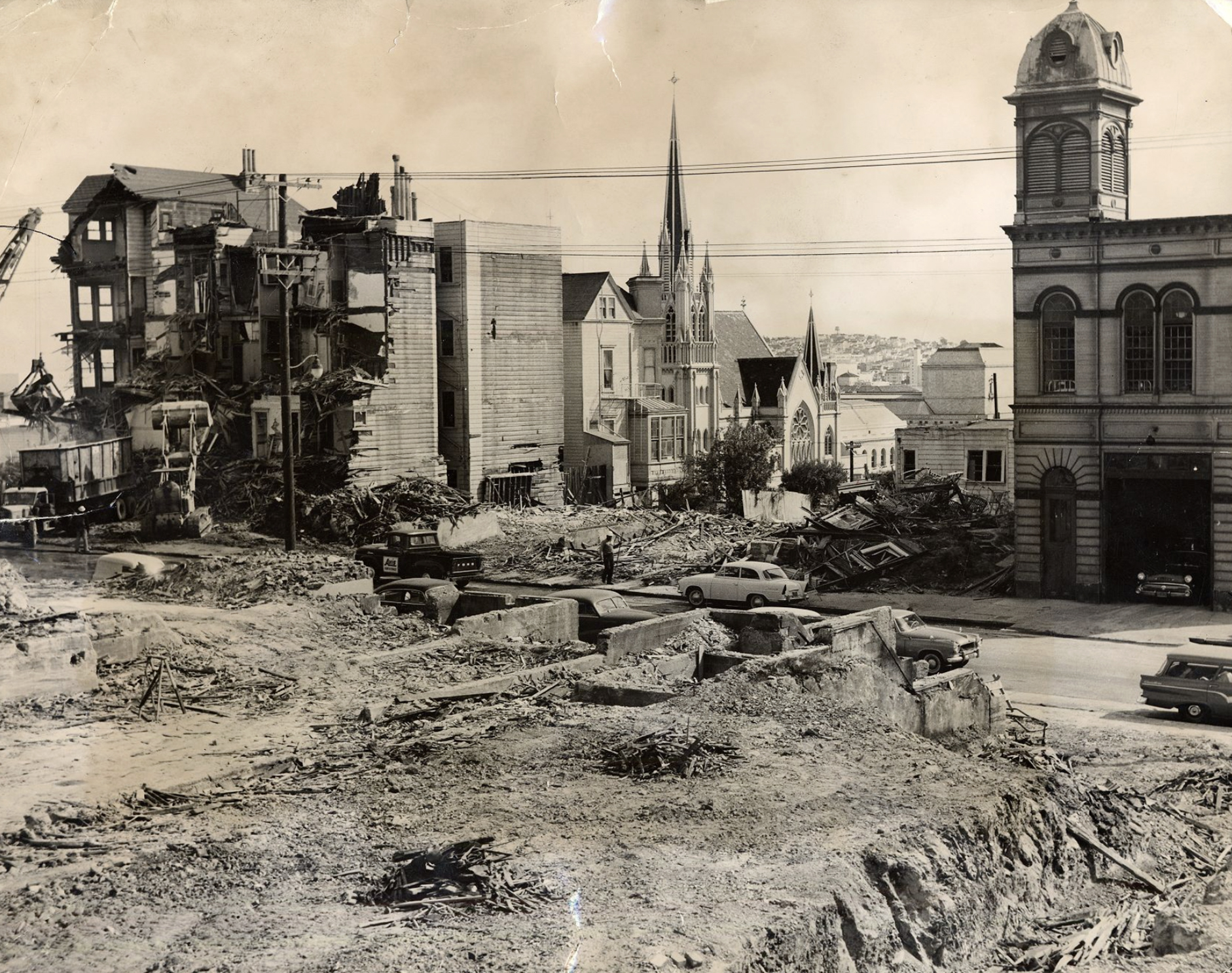 The image shows a cityscape with demolished buildings, debris, and construction vehicles. A church with pointed spires and an old building with a clock tower are visible.