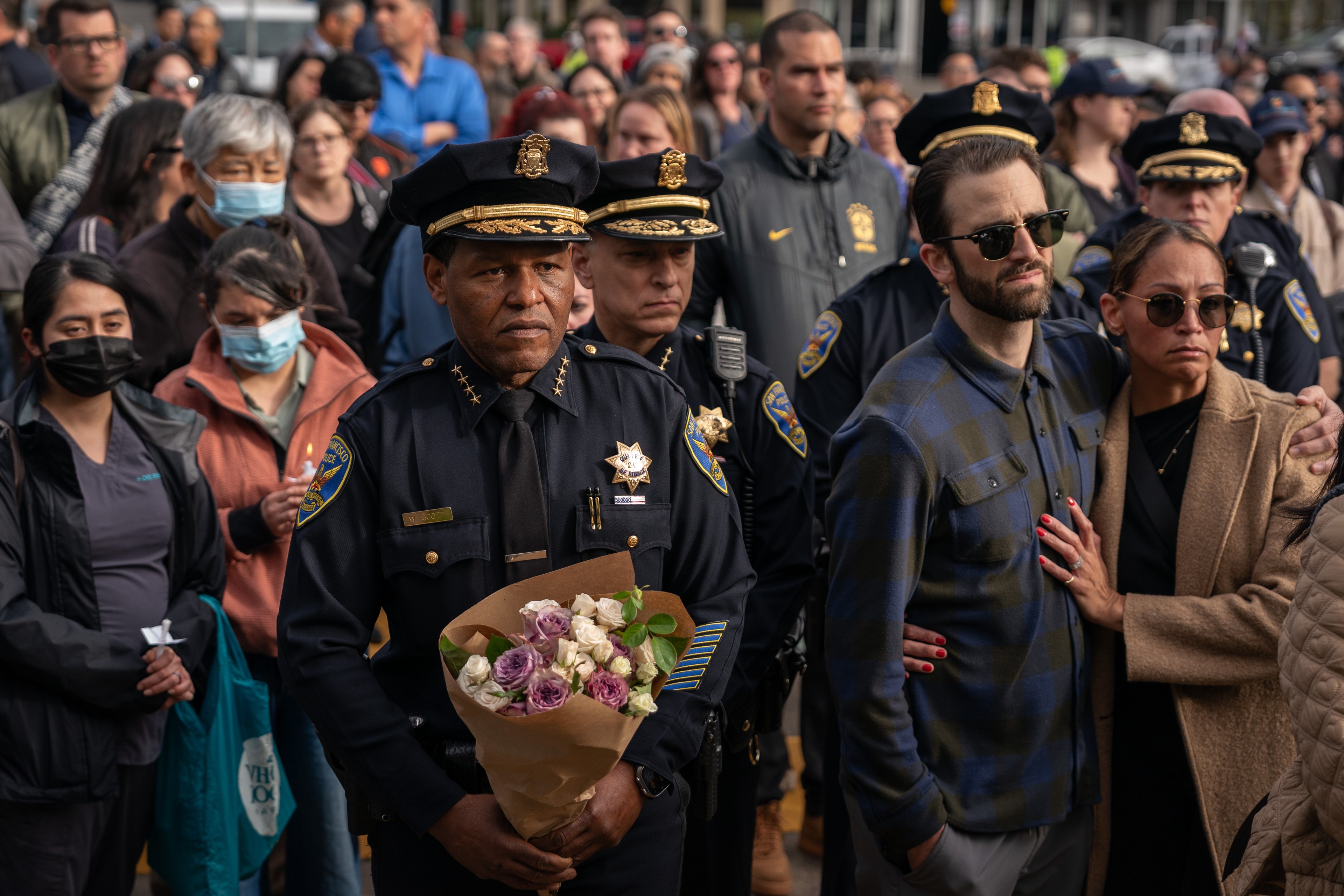 A solemn crowd stands together, with police officers in uniform at the front, one holding a bouquet of flowers. People appear serious and attentive.