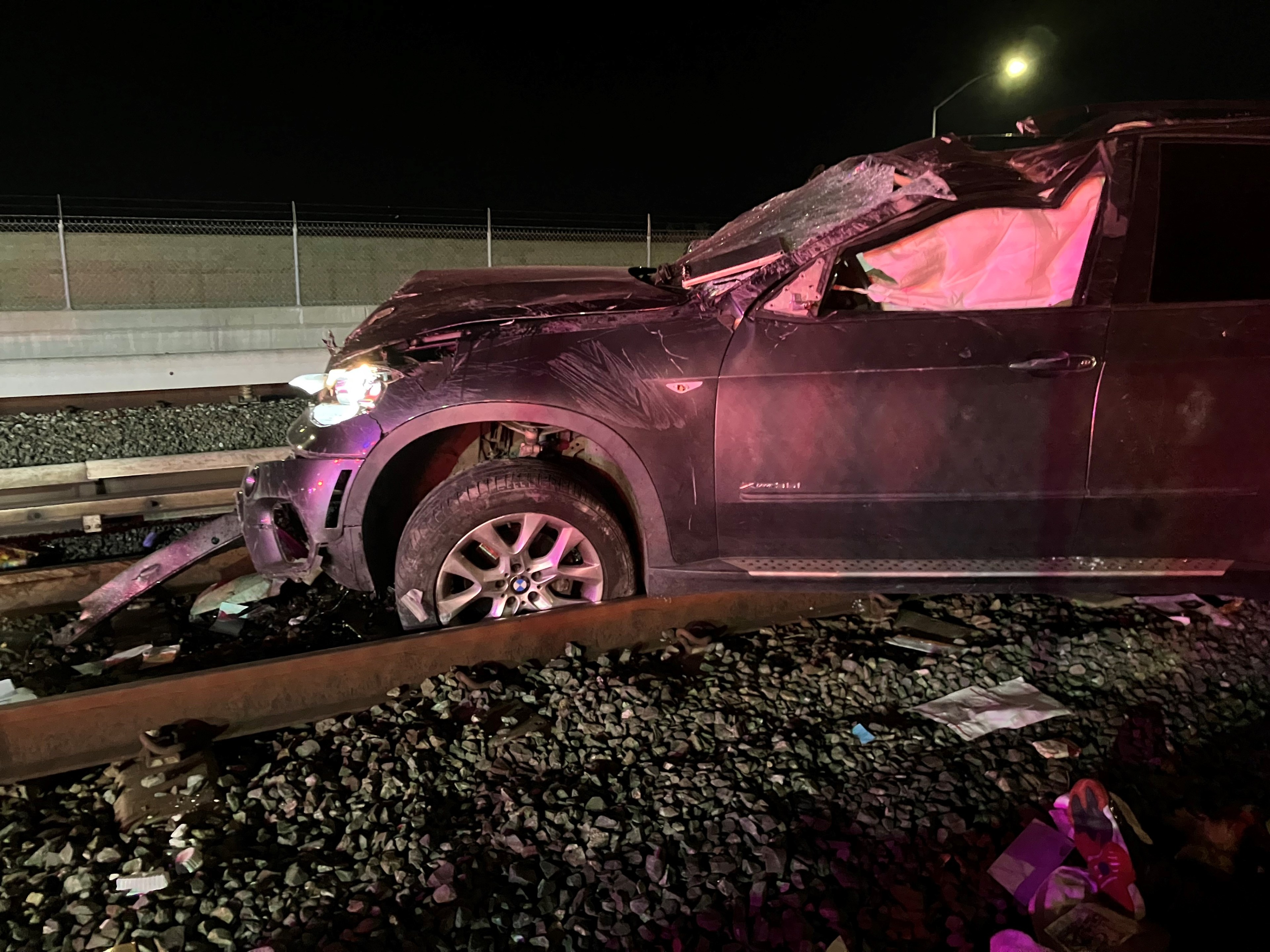 A damaged SUV is on railroad tracks at night, with a crumpled hood and shattered windshield. Debris is scattered around on the gravel.