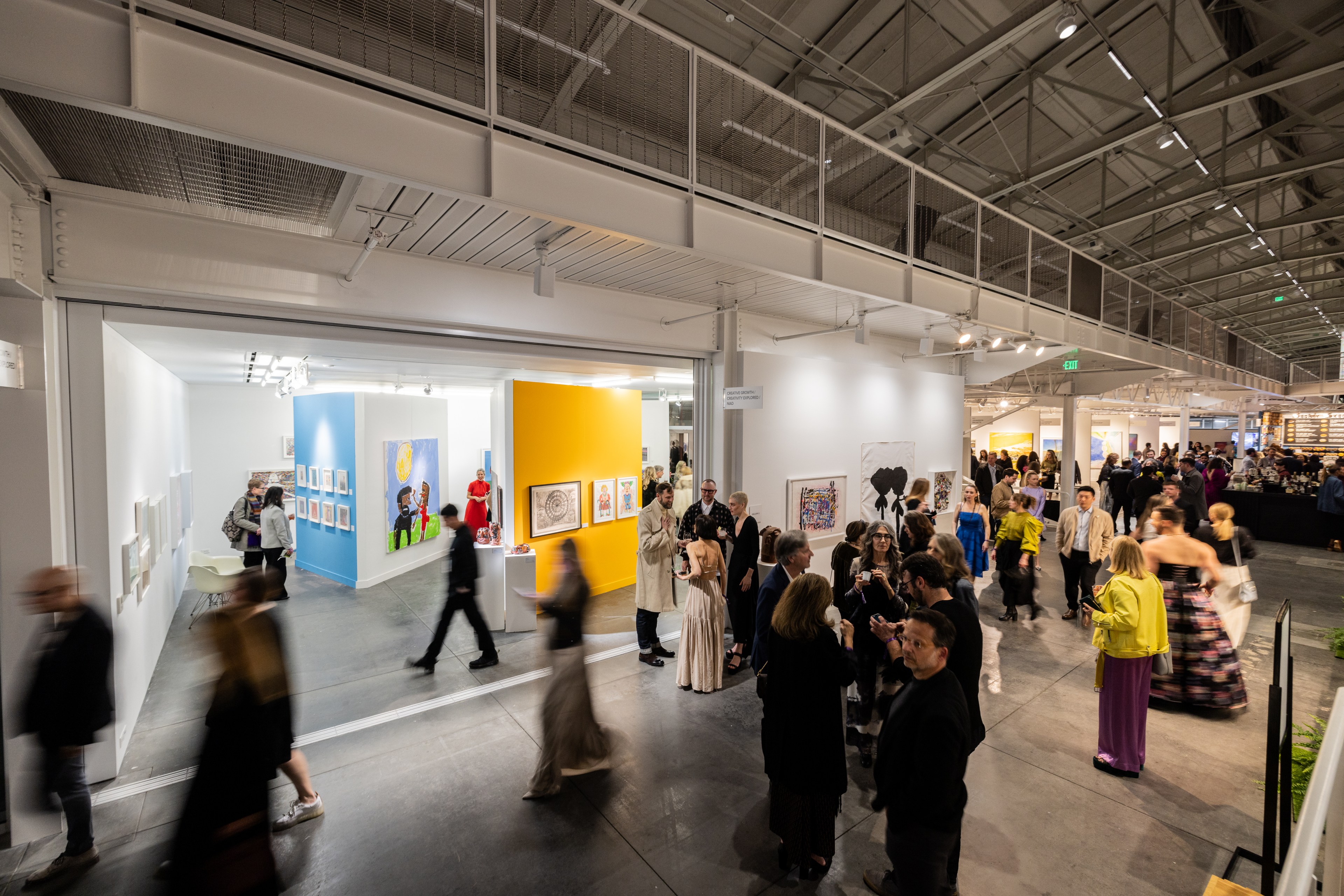 The image shows a busy art gallery with people walking and gathering. Vibrant artwork is displayed on walls, and the room has high ceilings and bright lighting.