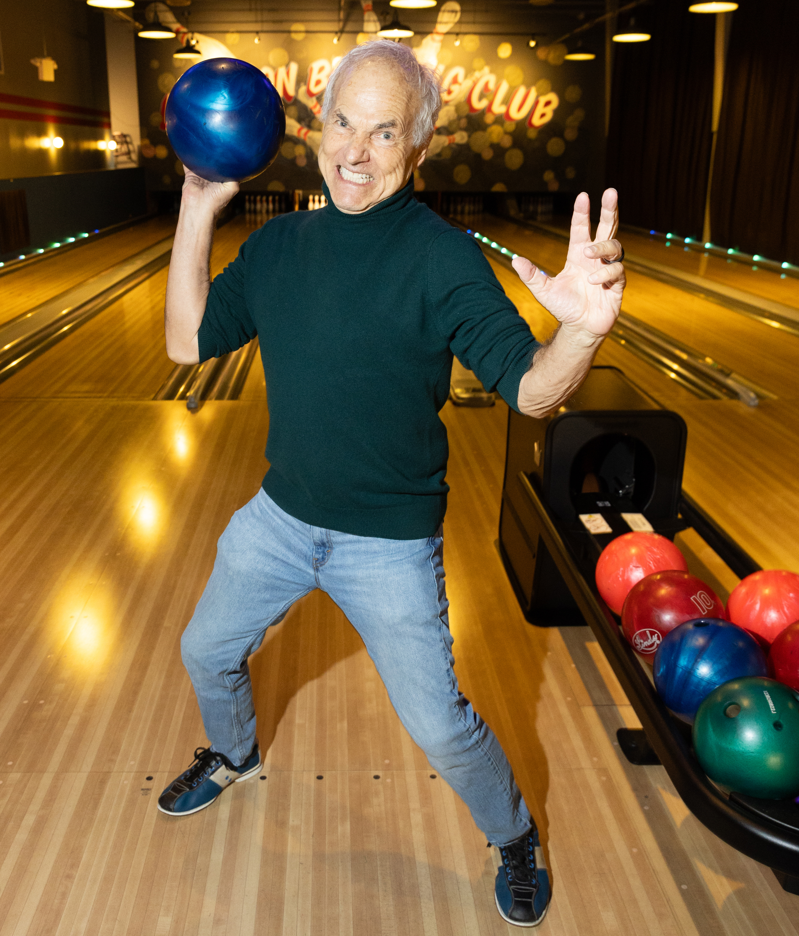 A person with gray hair is excitedly holding a blue bowling ball on a wooden lane, with colorful bowling balls in a rack beside them.