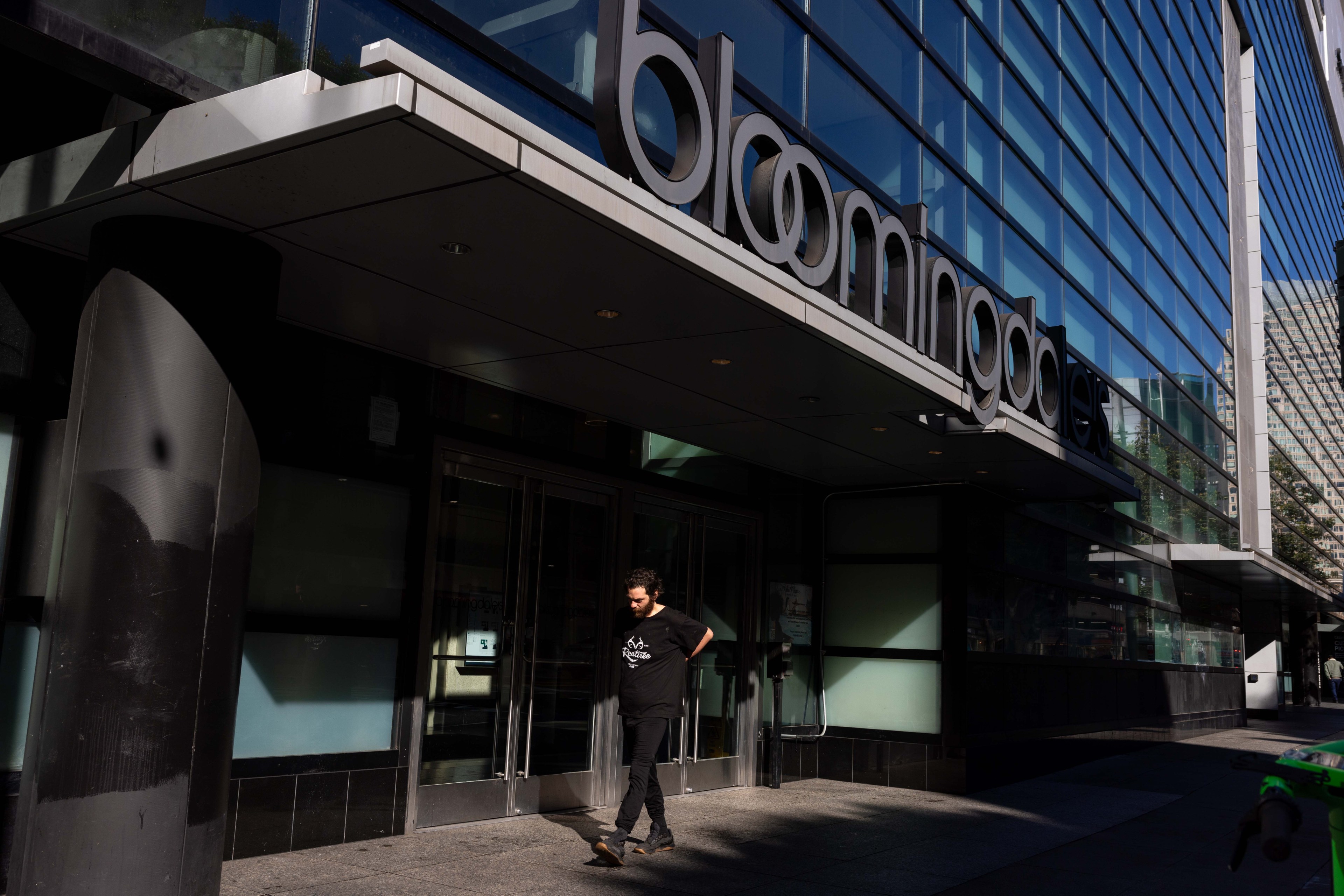 A person walks in front of a Bloomingdale's store, with a modern glass and metal facade. Sunlight casts shadows on the pavement and building entrance.