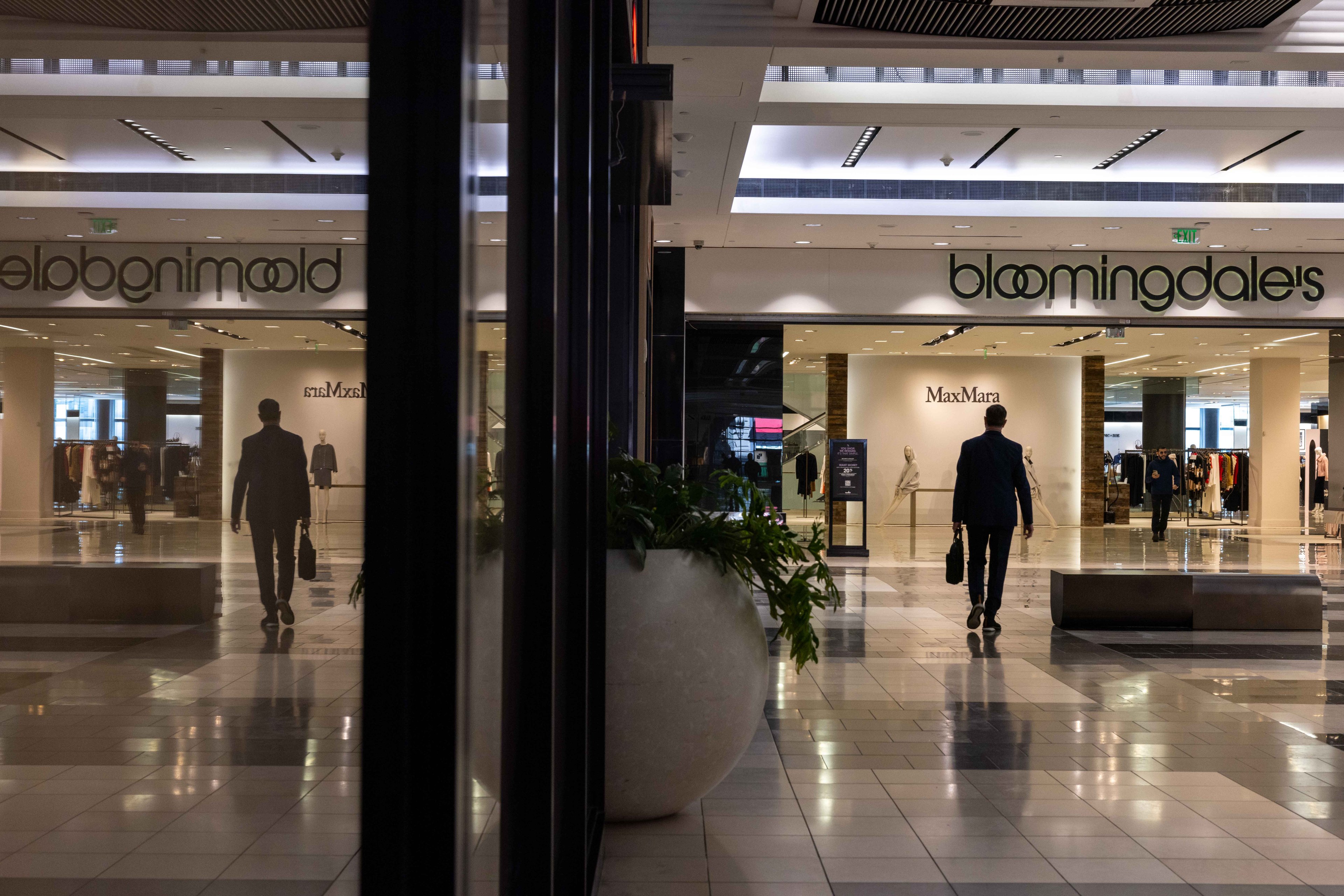 A man walks inside a glossy mall toward Bloomingdale's and Max Mara, with his reflection visible on the polished floor and a potted plant nearby.