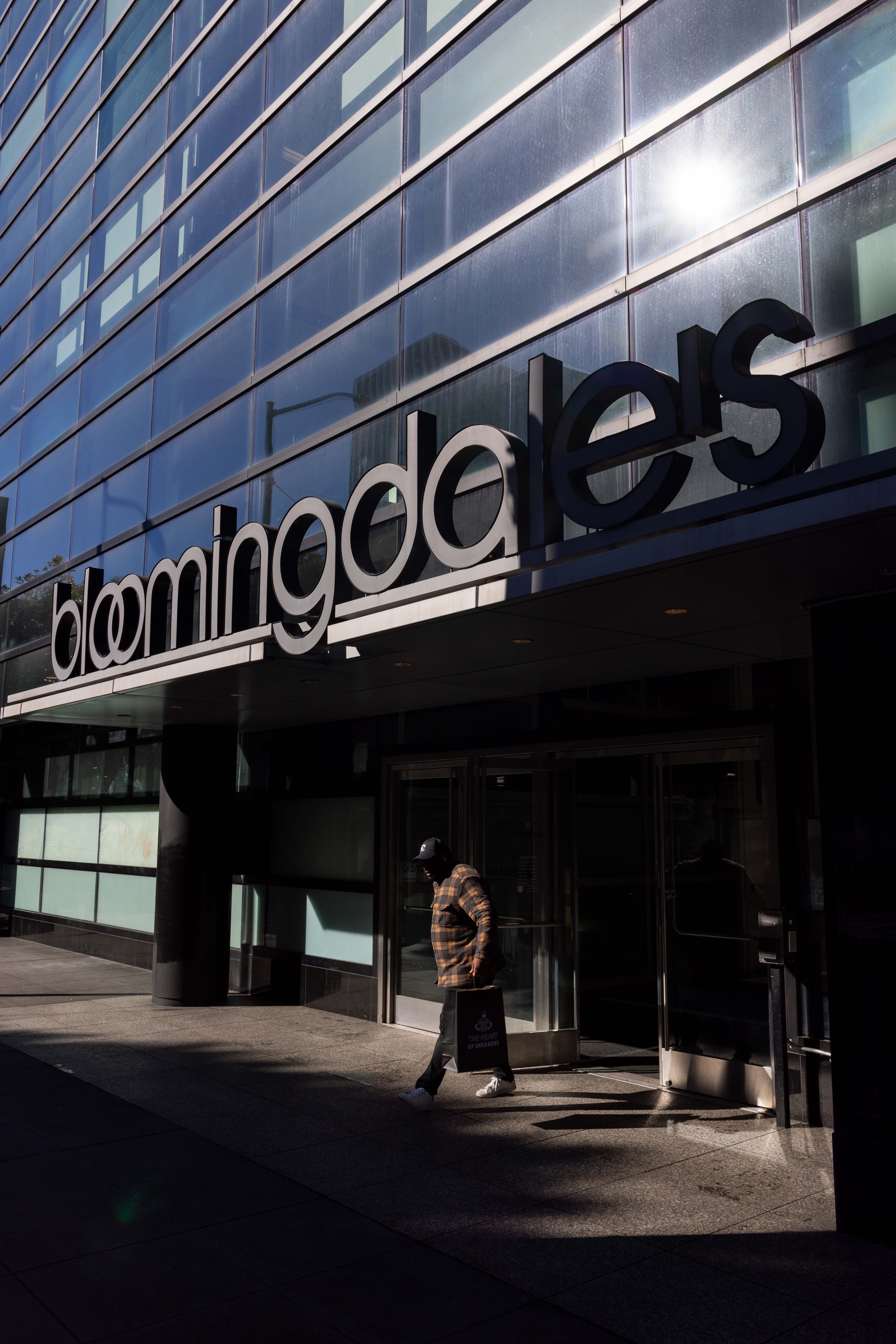 A person walks out of a Bloomingdale's store, carrying a shopping bag. The building has large, reflective windows and the sun shines brightly.