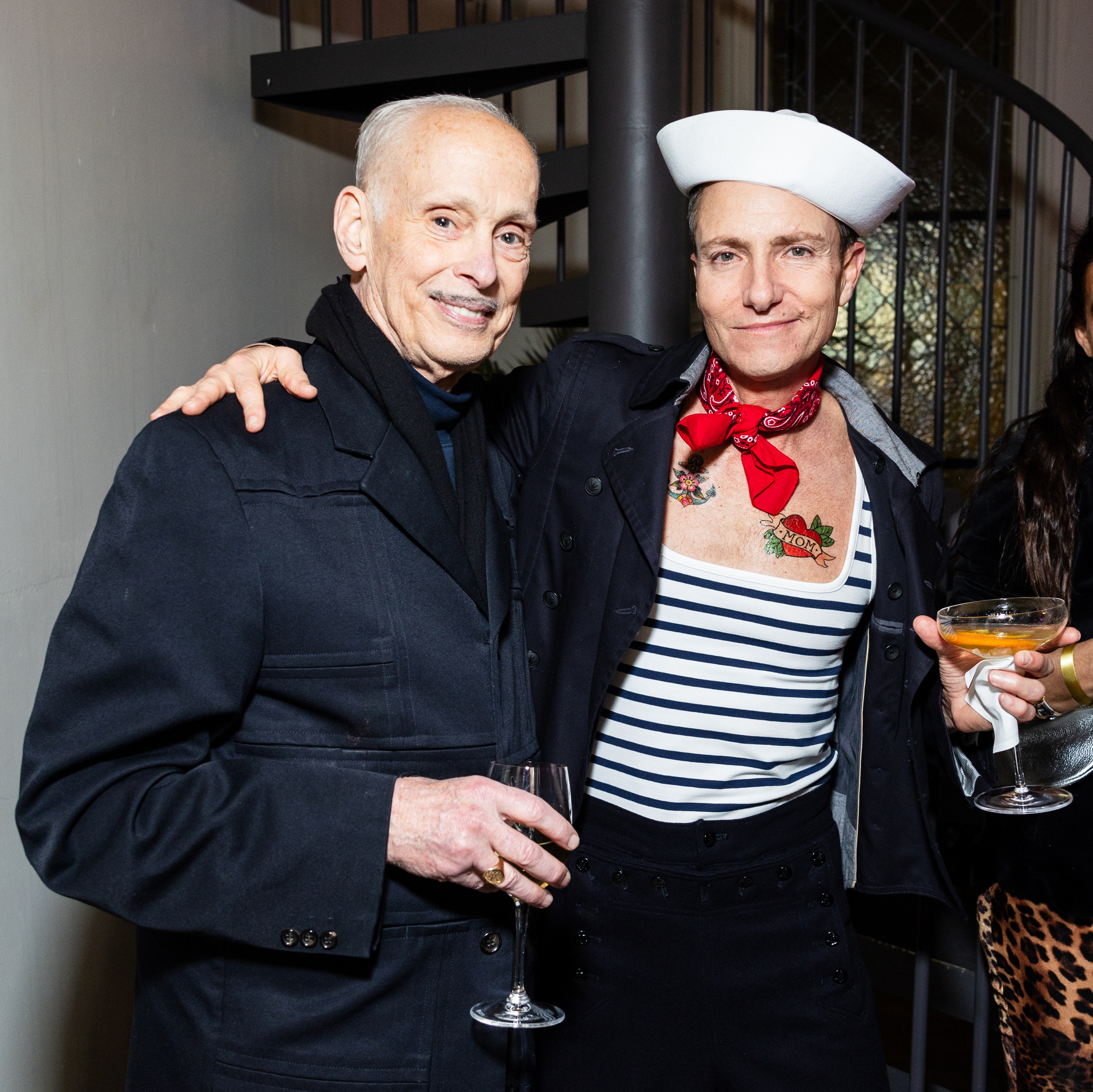 Two people pose together. One wears a dark coat and holds a wine glass, and the other is dressed in a sailor outfit with a cocktail. They appear to be at a social event.