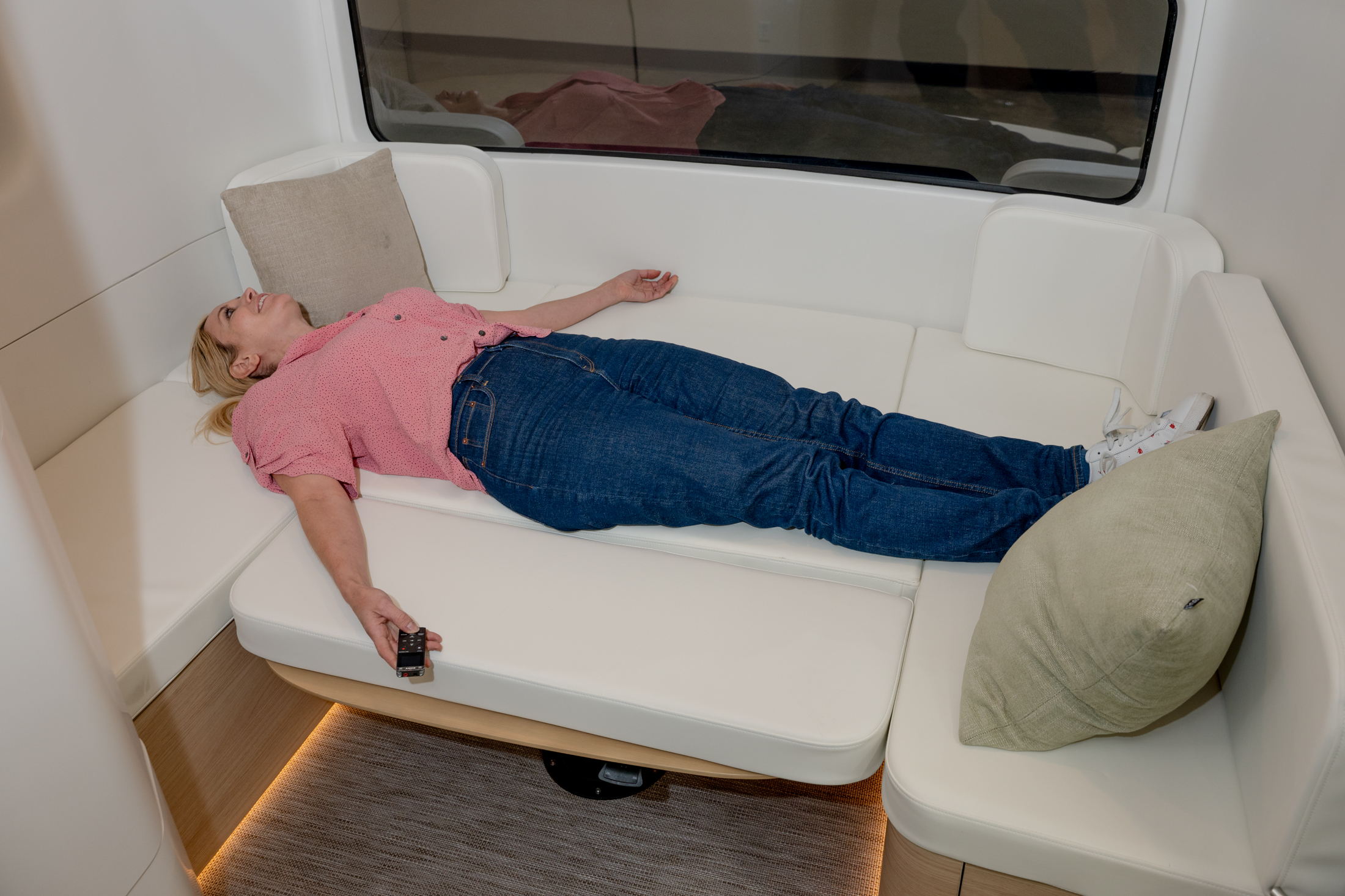 A person in a pink shirt and jeans lies on a white L-shaped couch holding a remote, with a beige pillow beside them and soft lighting underneath.