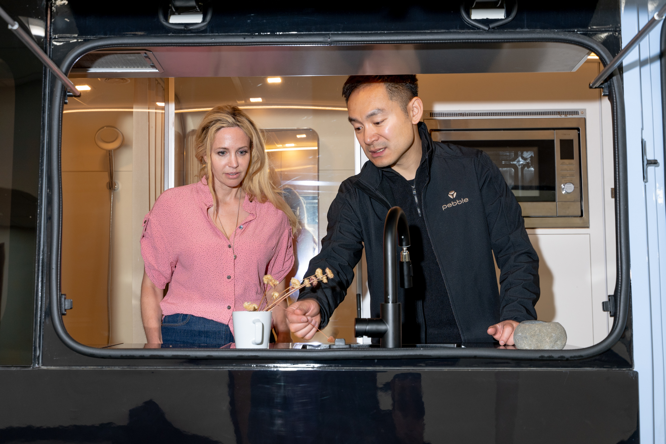 A woman in a pink shirt and a man in a black jacket stand in a modern kitchen. The man gestures towards a mug with dried flowers, behind a sleek sink.