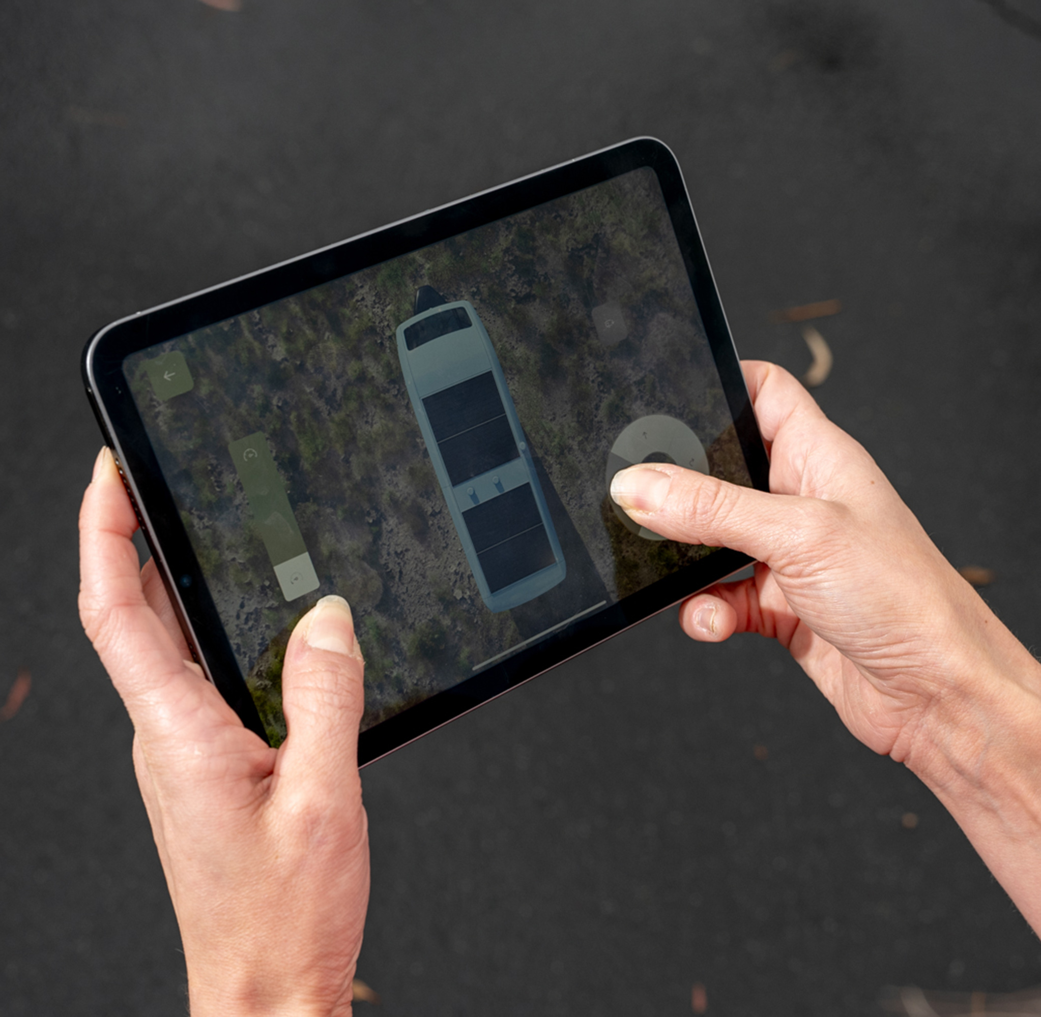 Two hands are holding a tablet showing an aerial view of a vehicle on a rough terrain. The screen displays controls and navigation features.