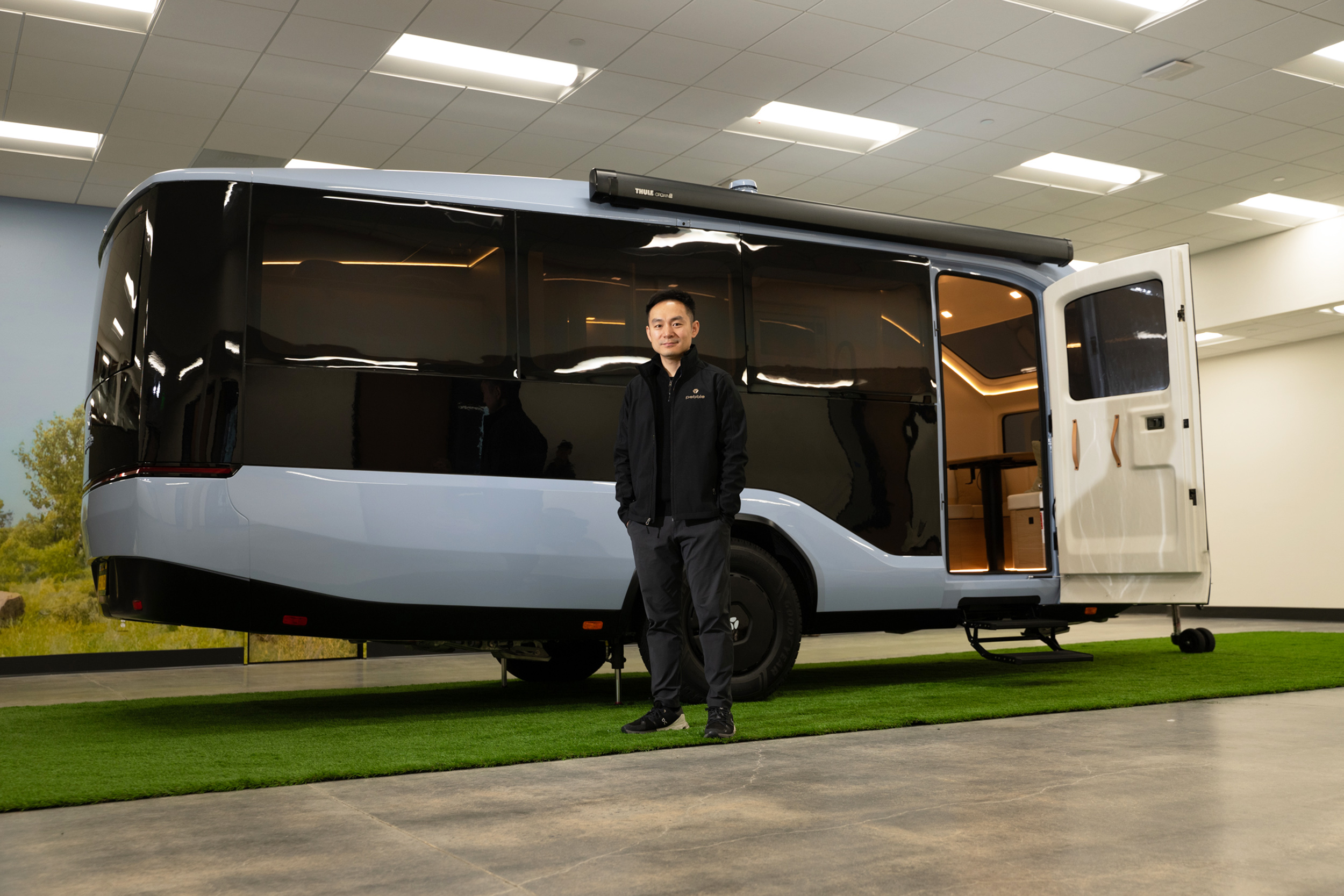 A man stands in front of a sleek, modern camper trailer with large black tinted windows and an open door, displayed indoors on a patch of green grass.