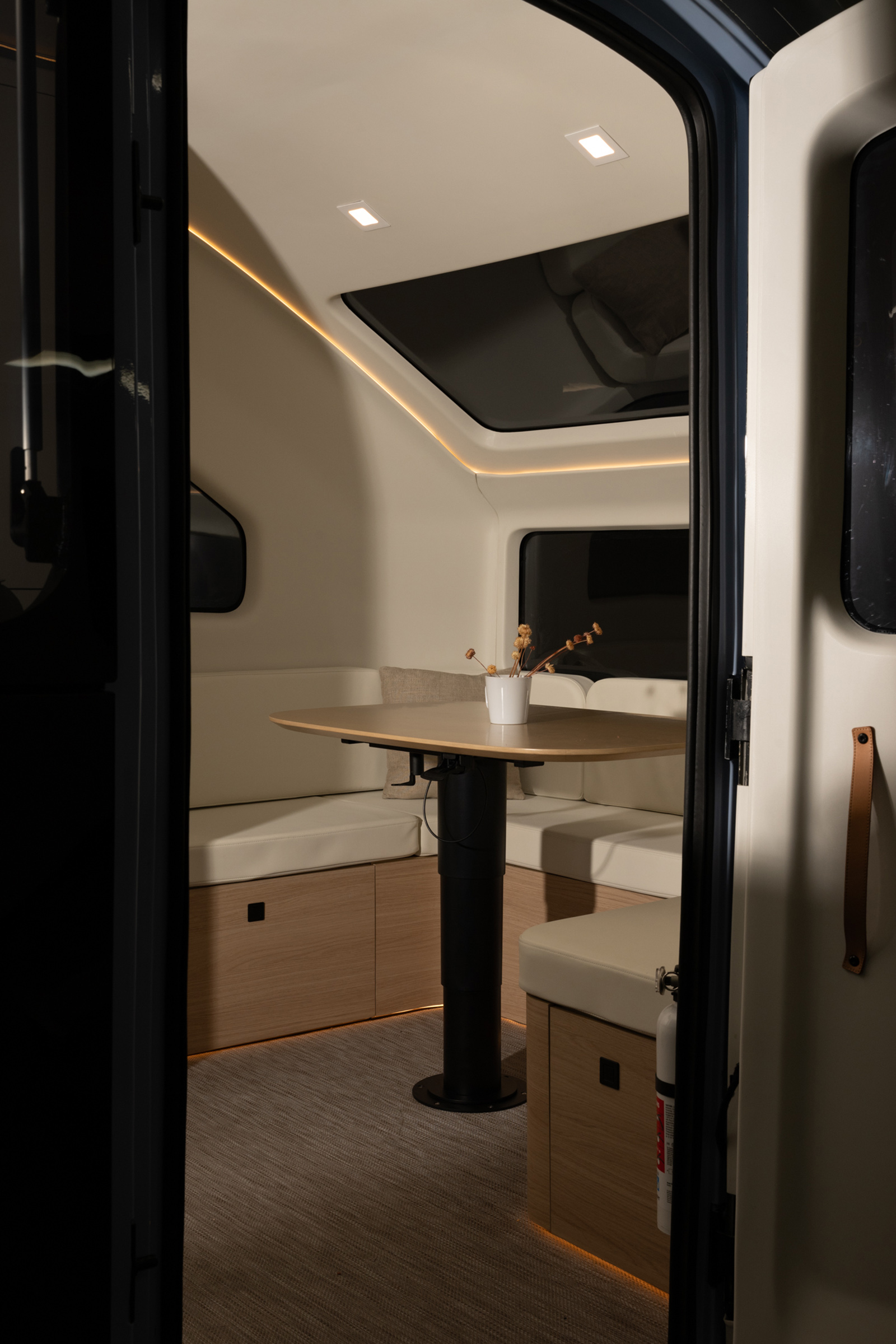 A cozy RV interior with a skylight displays a cushioned seating area around a small table with a potted plant, featuring sleek lighting and wooden accents.