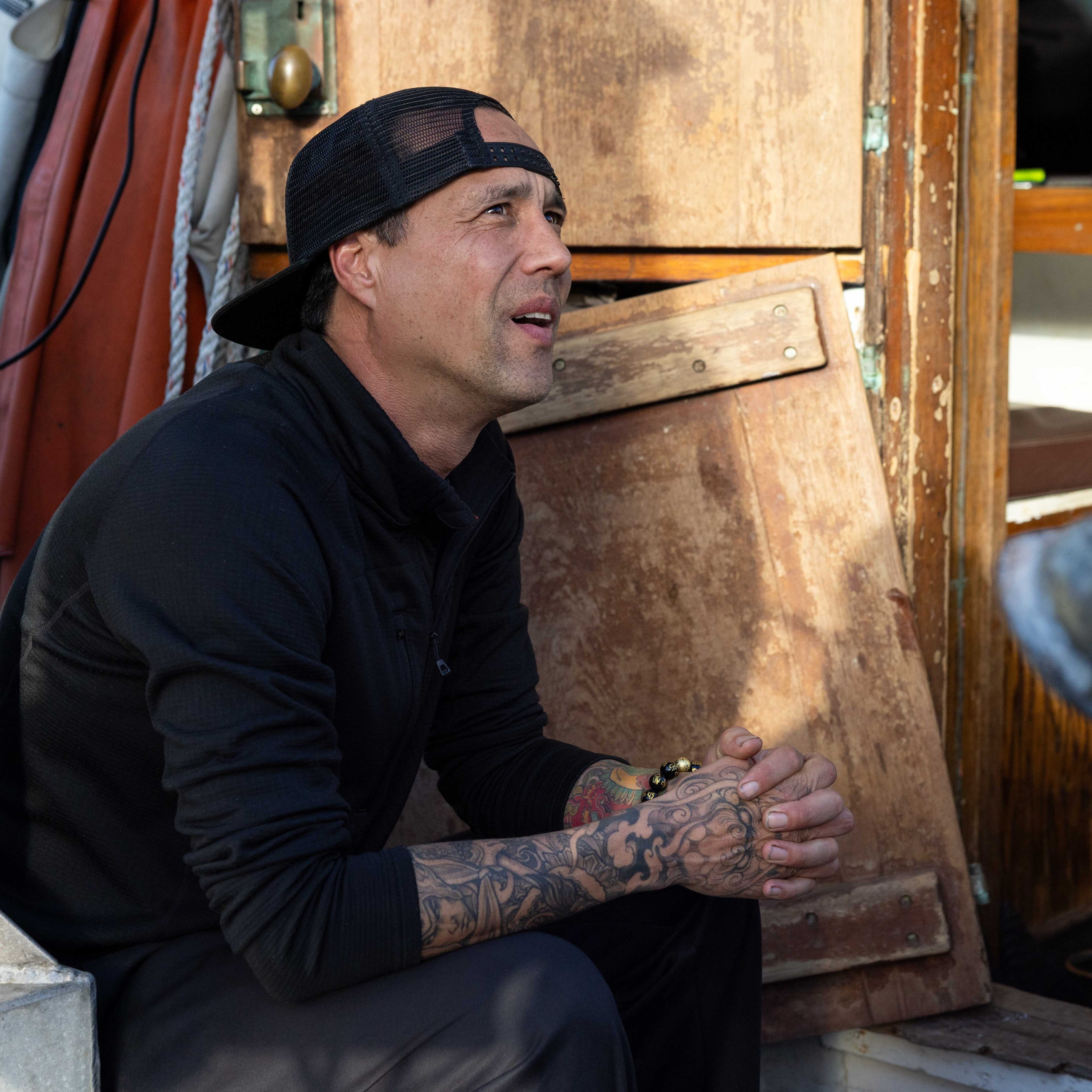 A person with tattoos and a black cap worn backward sits thoughtfully in a boat. The surroundings feature ropes, wooden panels, and light casting shadows.