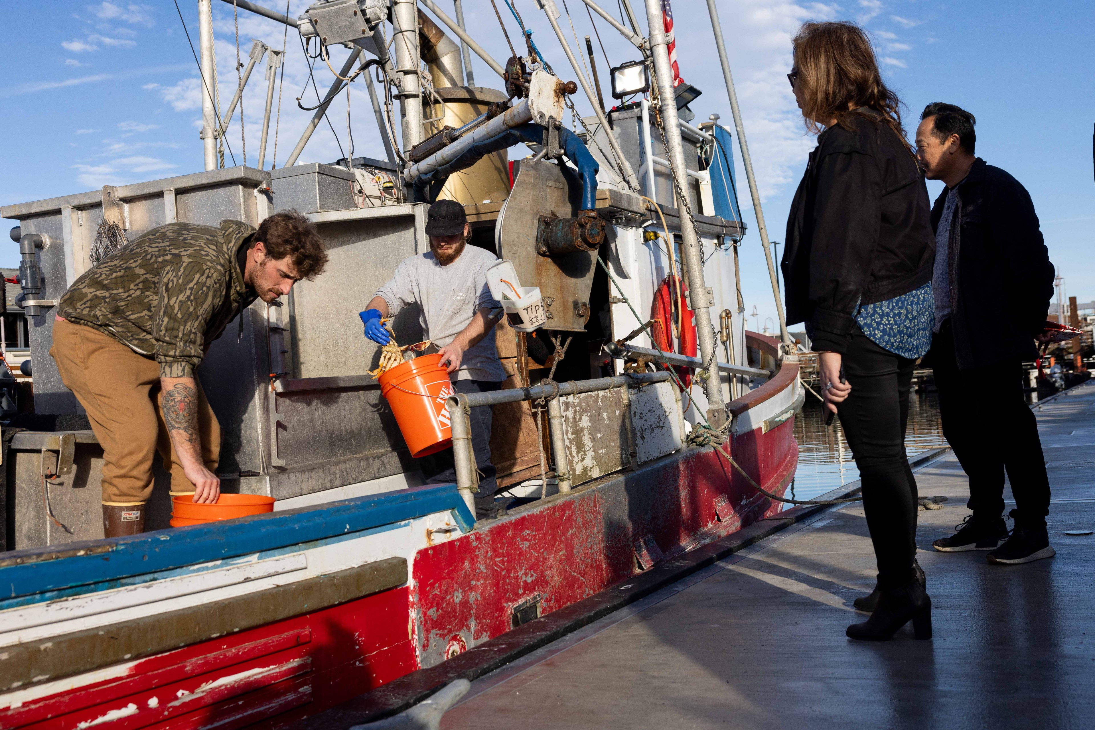 Two men on a fishing boat handle orange buckets, while two people stand on the dock watching. The boat has a weathered, industrial look under a blue sky.