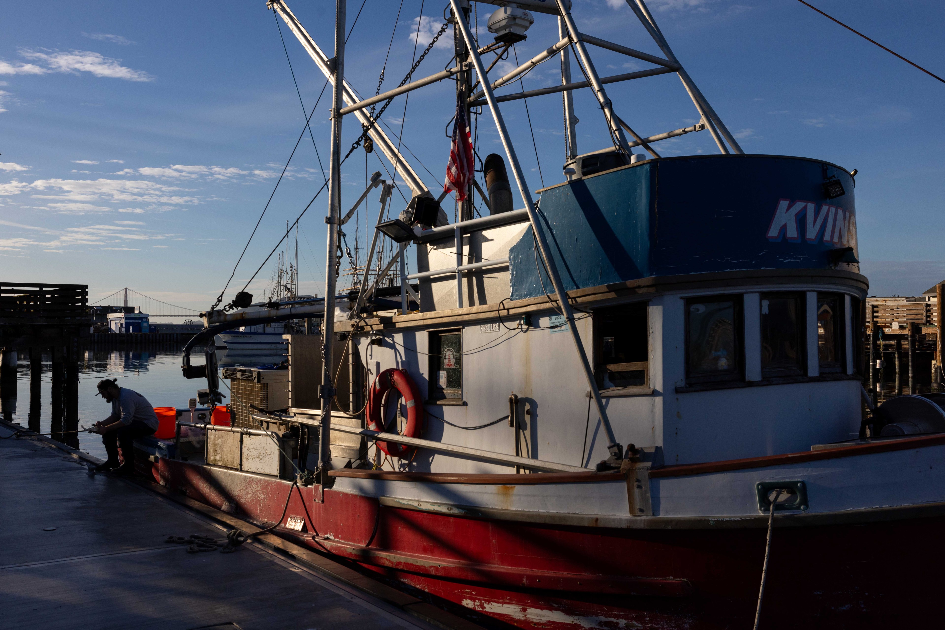 A fishing boat is docked at a pier, with a person working beside it. The boat has a blue and red hull, and there's an American flag on the mast.