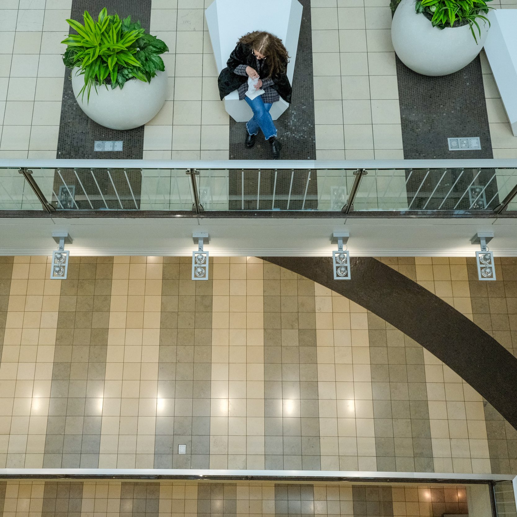 A person sits on a white bench next to large potted plants on tiled flooring, viewed from above. They appear to be looking at something in their hands.