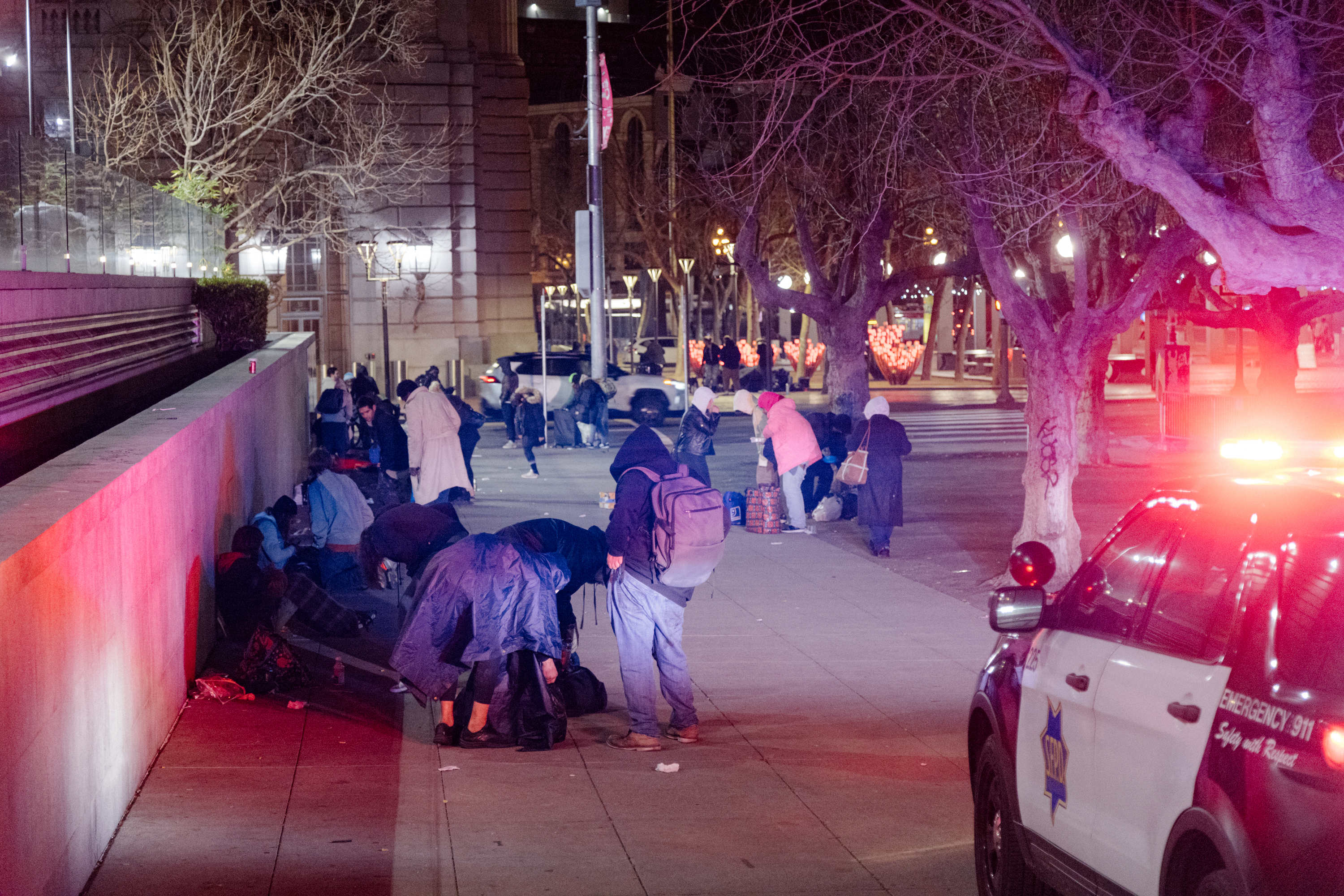 A group of people is gathered at night on a city sidewalk, with a police car parked nearby. Many are wearing coats and appear to be outdoors in cold weather.