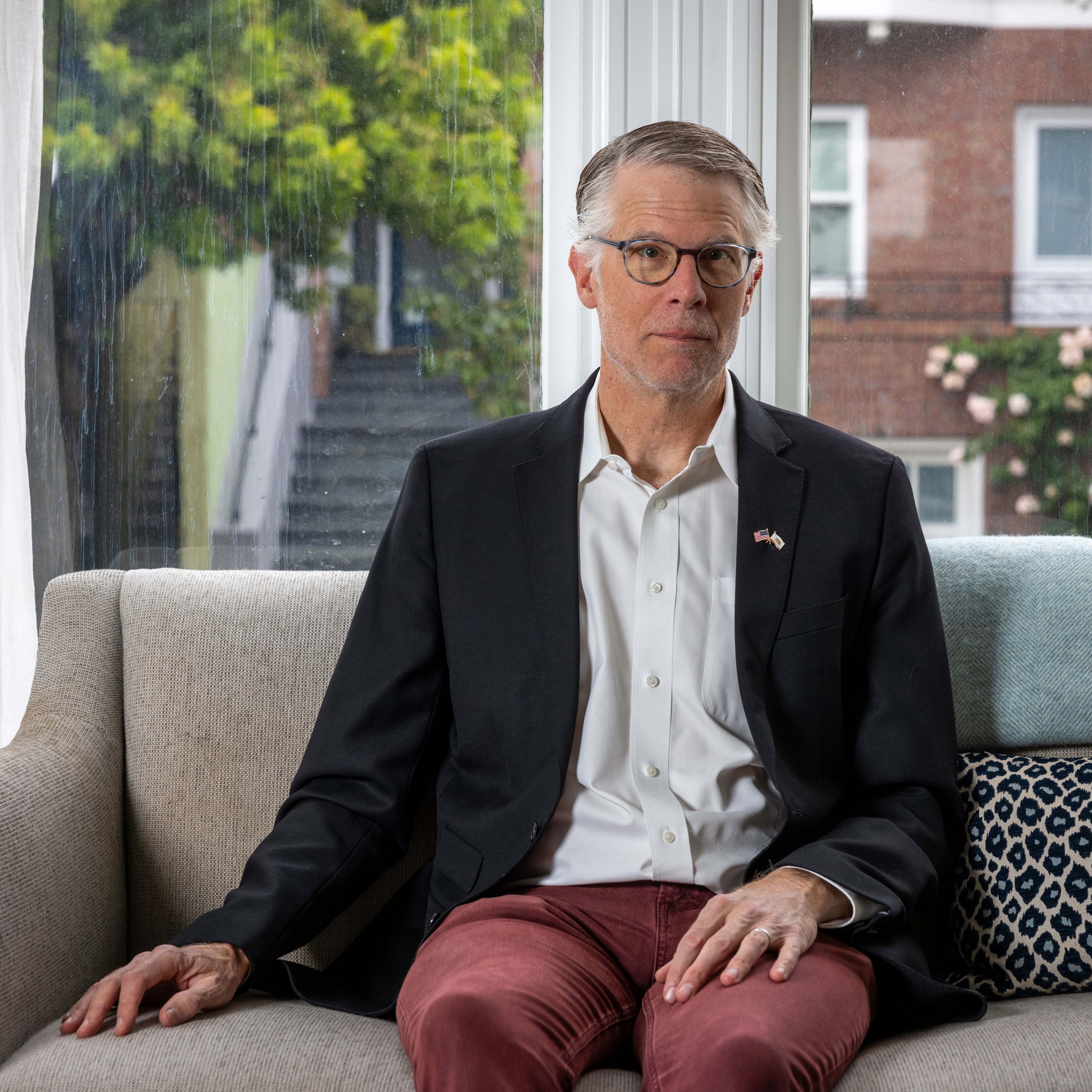 A man with glasses is seated on a beige sofa. He wears a black suit jacket, white shirt, and red pants. A window with greenery and a brick building is behind him.