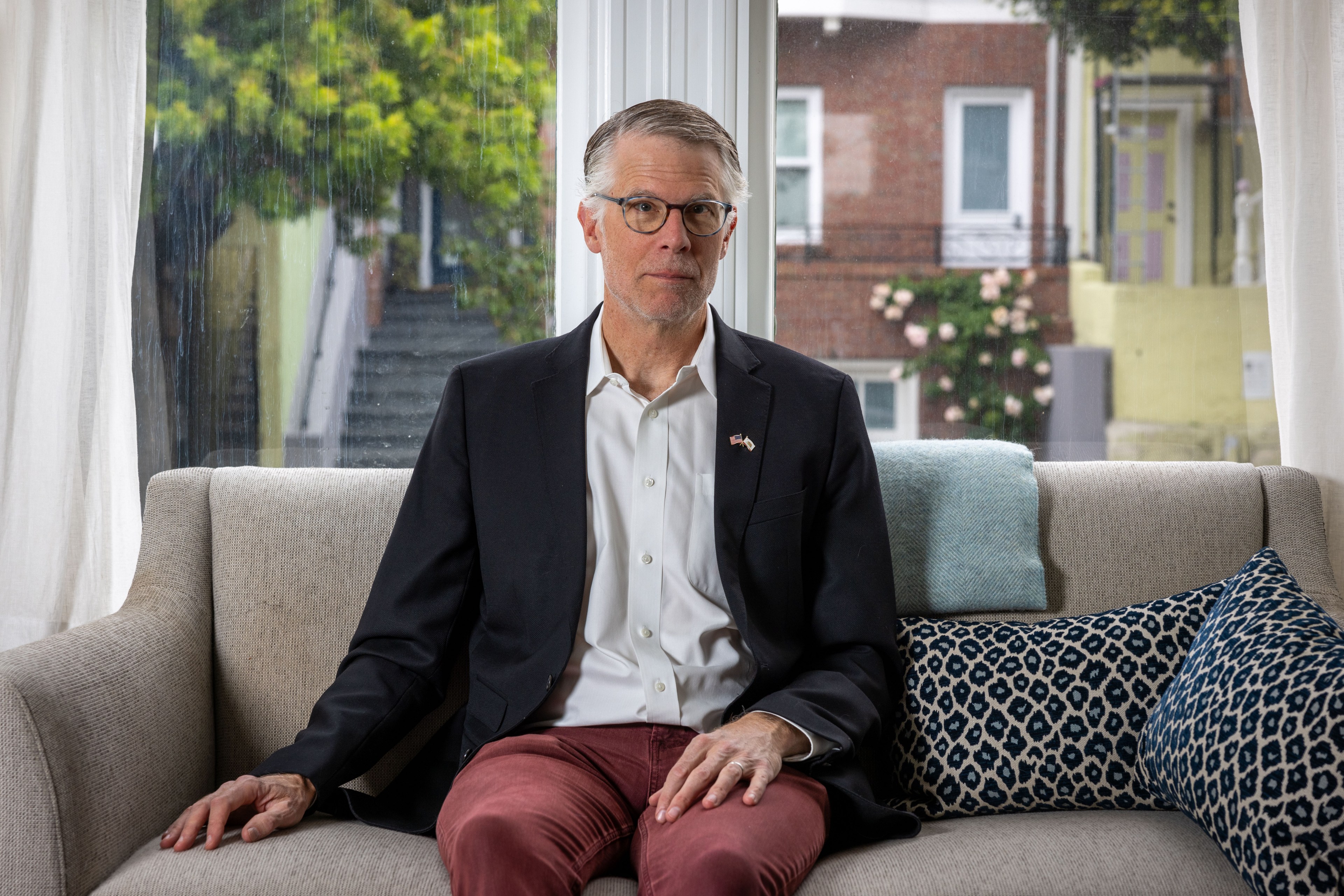 A man in a suit and glasses sits on a beige sofa with patterned cushions, against a backdrop of a window showing greenery and houses outside.