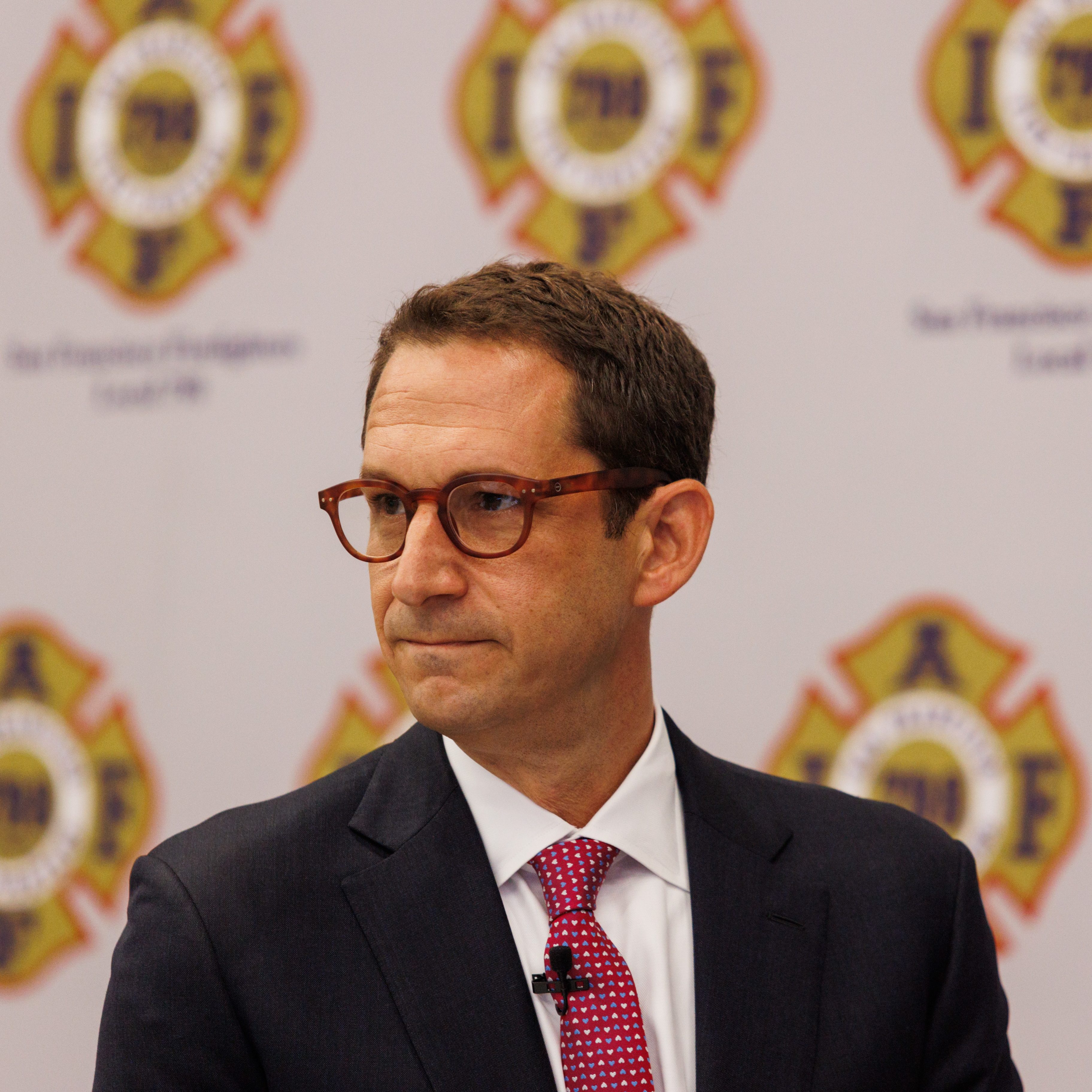 A man in a suit and red tie stands in front of a backdrop with fire department logos, looking to the side. The logos read "San Francisco Firefighters Local 798."