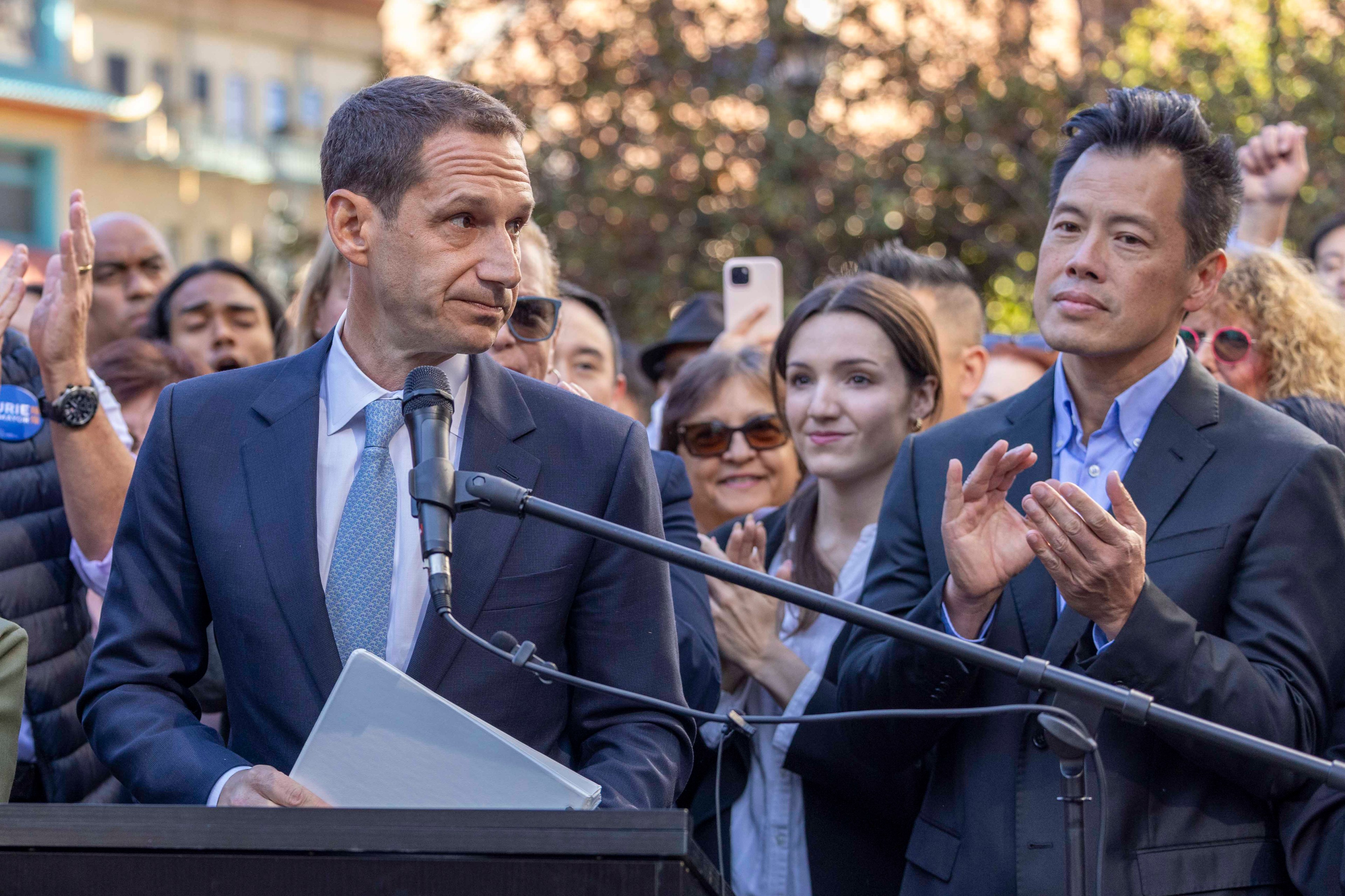 A man in a suit speaks at a podium with a microphone, holding a folder. Another man in a suit applauds, while a crowd with phones and smiles observes.