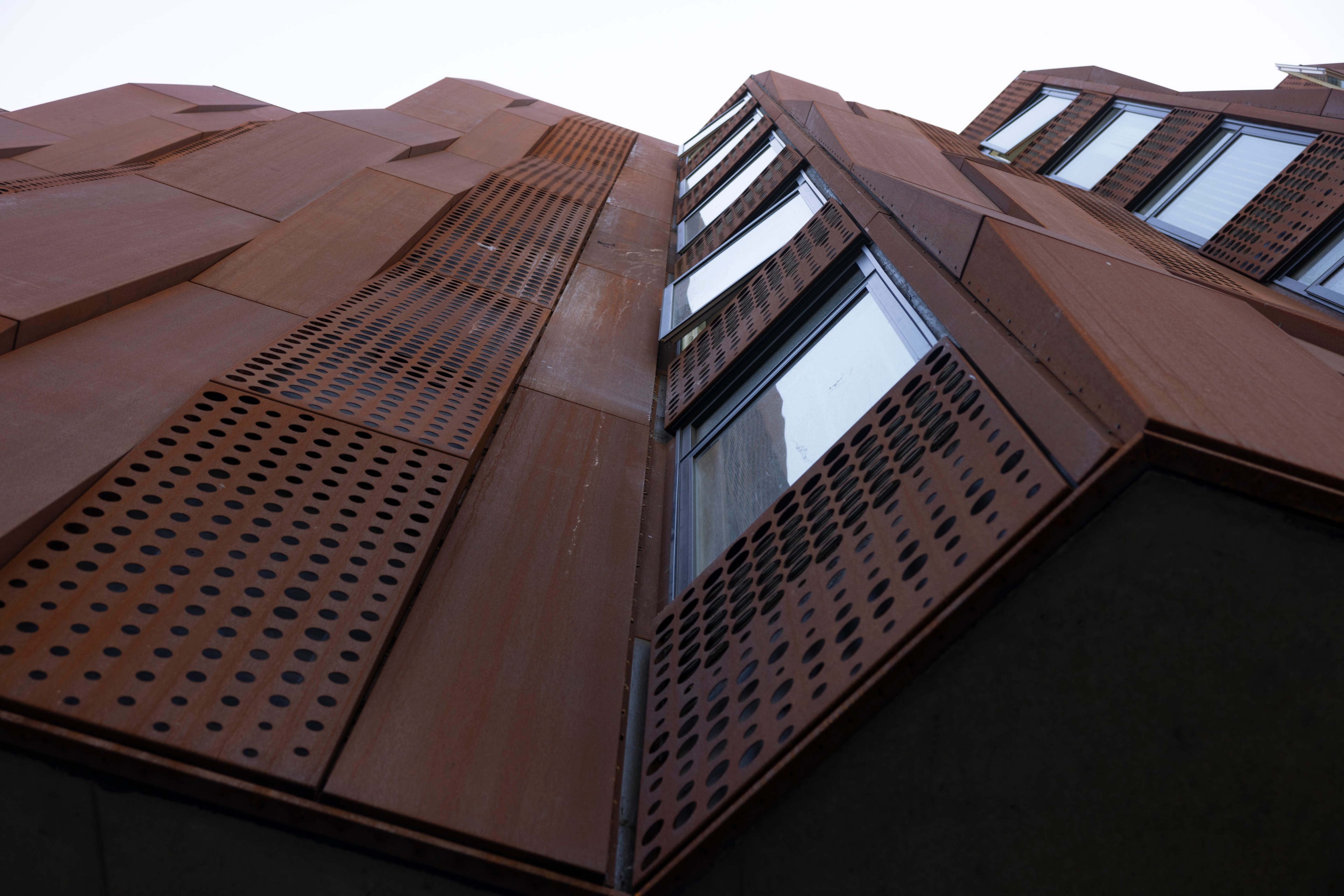 The image shows an upward view of a modern building facade with rust-colored panels and large windows, featuring a grid of circular perforations.