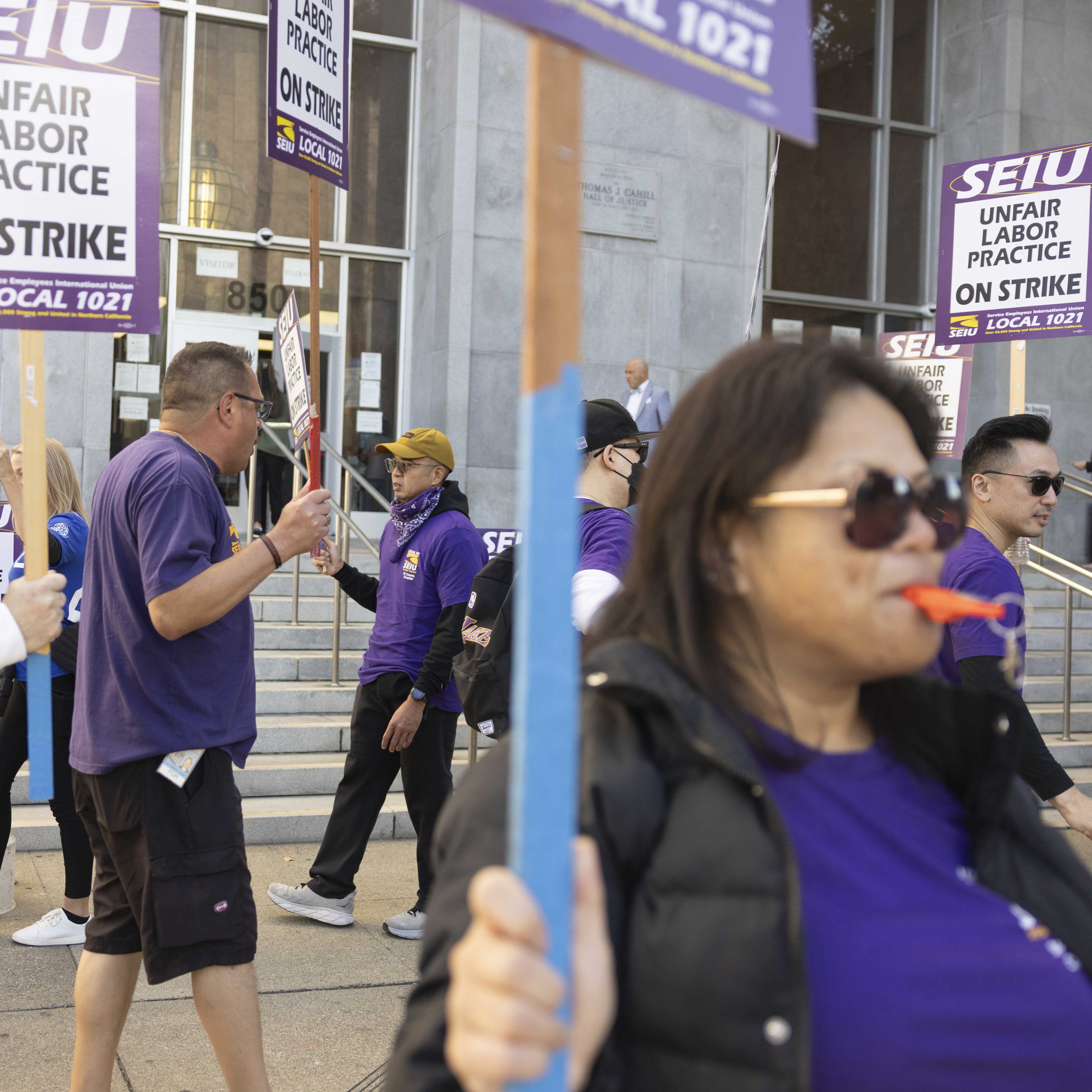 People in purple shirts holding &quot;Unfair Labor Practice On Strike&quot; signs are gathered in front of a building, participating in a strike or protest.