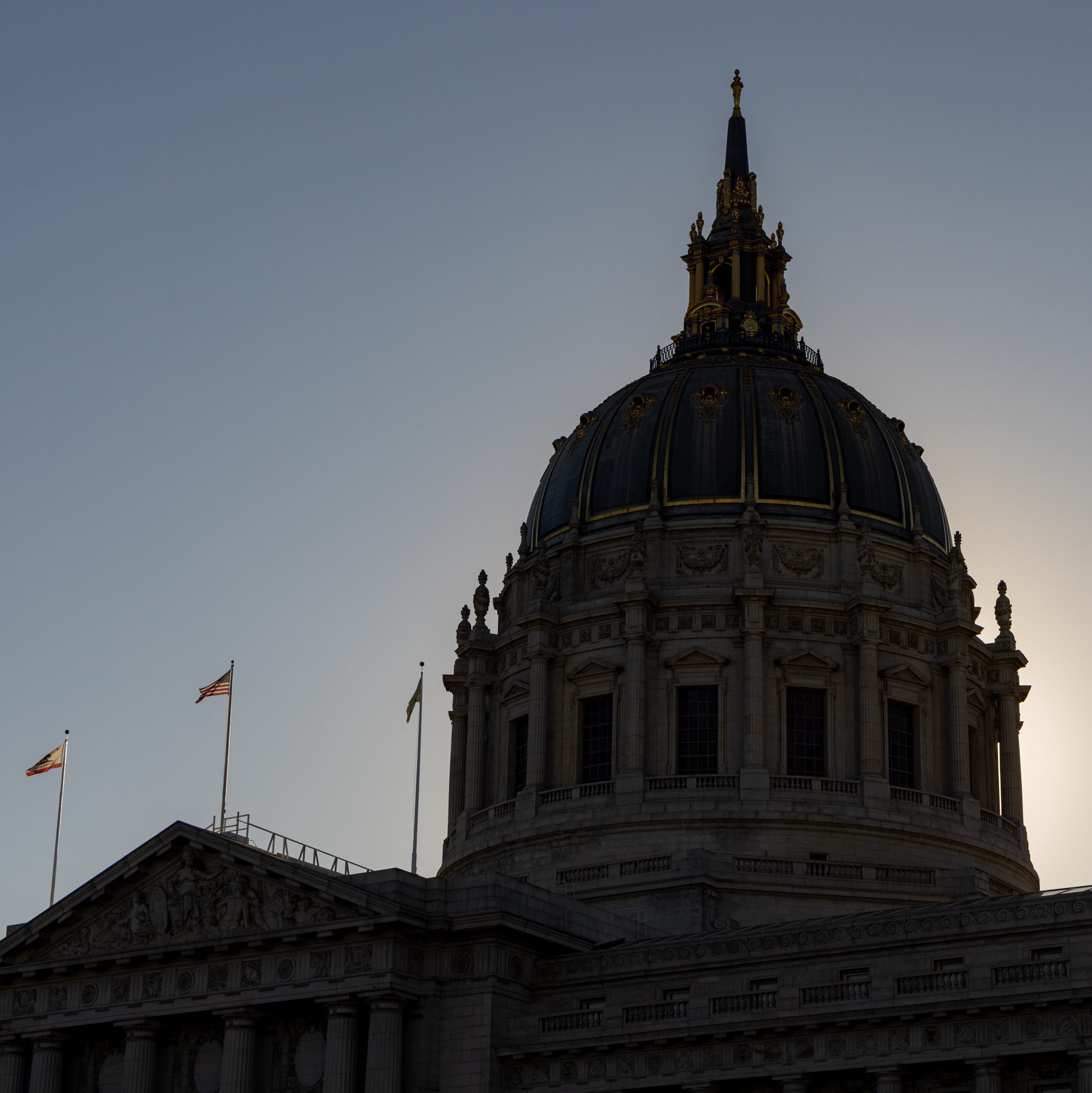 A grand building with a large domed roof, adorned with ornate details, stands against a clear sky. Two flags are visible on poles nearby.