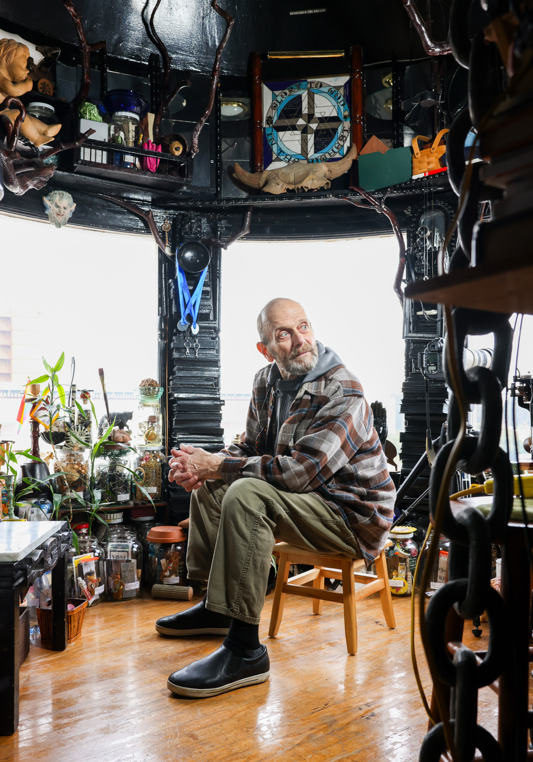 A bearded man sits on a wooden stool in a cluttered room filled with plants, jars, and eclectic decor. He wears a plaid shirt, green pants, and black shoes.