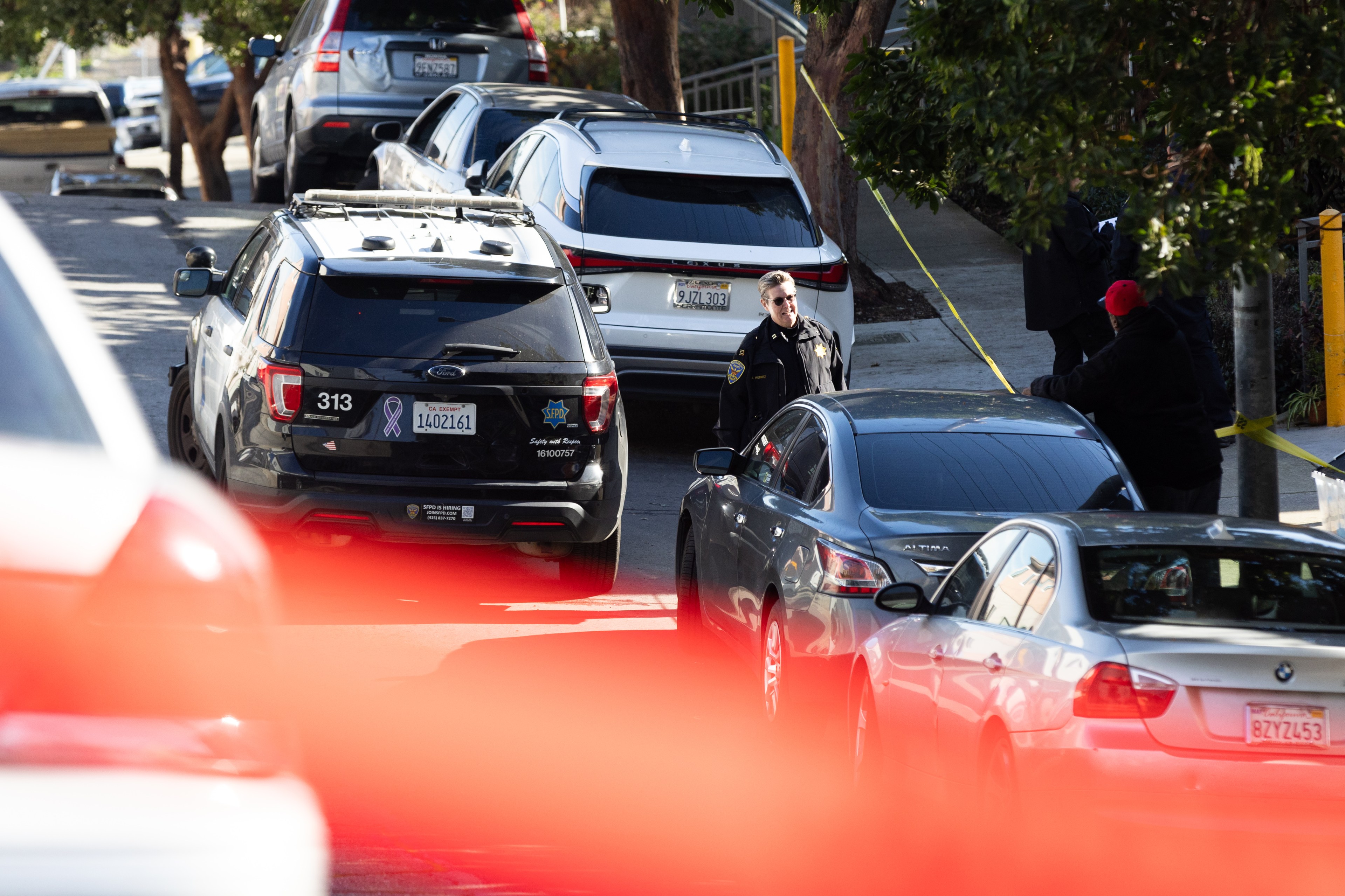 The image shows a street scene with several parked cars, a police SUV, and two people, one in police uniform, standing near caution tape. Red foreground blur is visible.