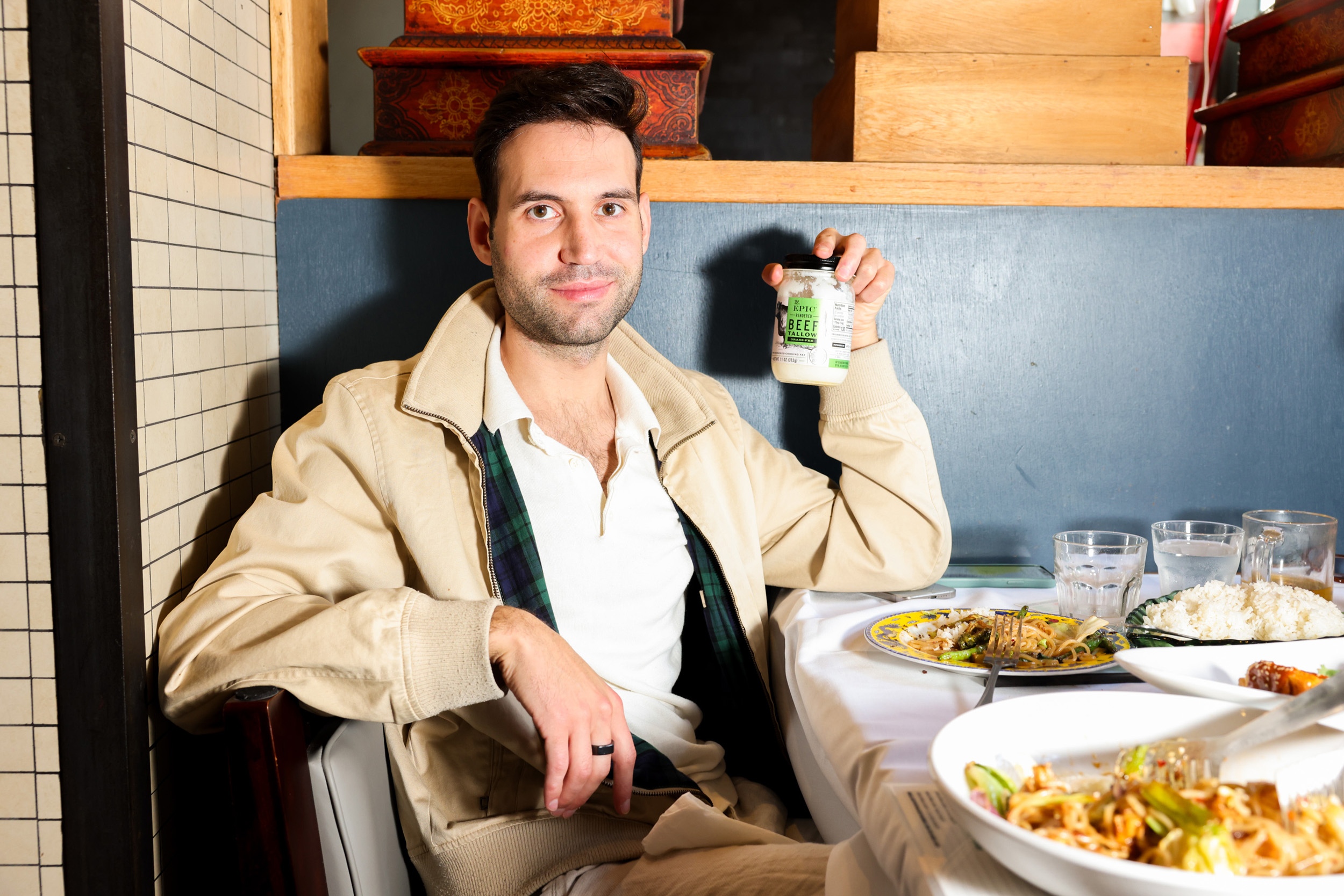 A man sits in a restaurant holding a jar labeled "Beef" with a half-smile. There are plates of food and glasses of water on the table in front of him.