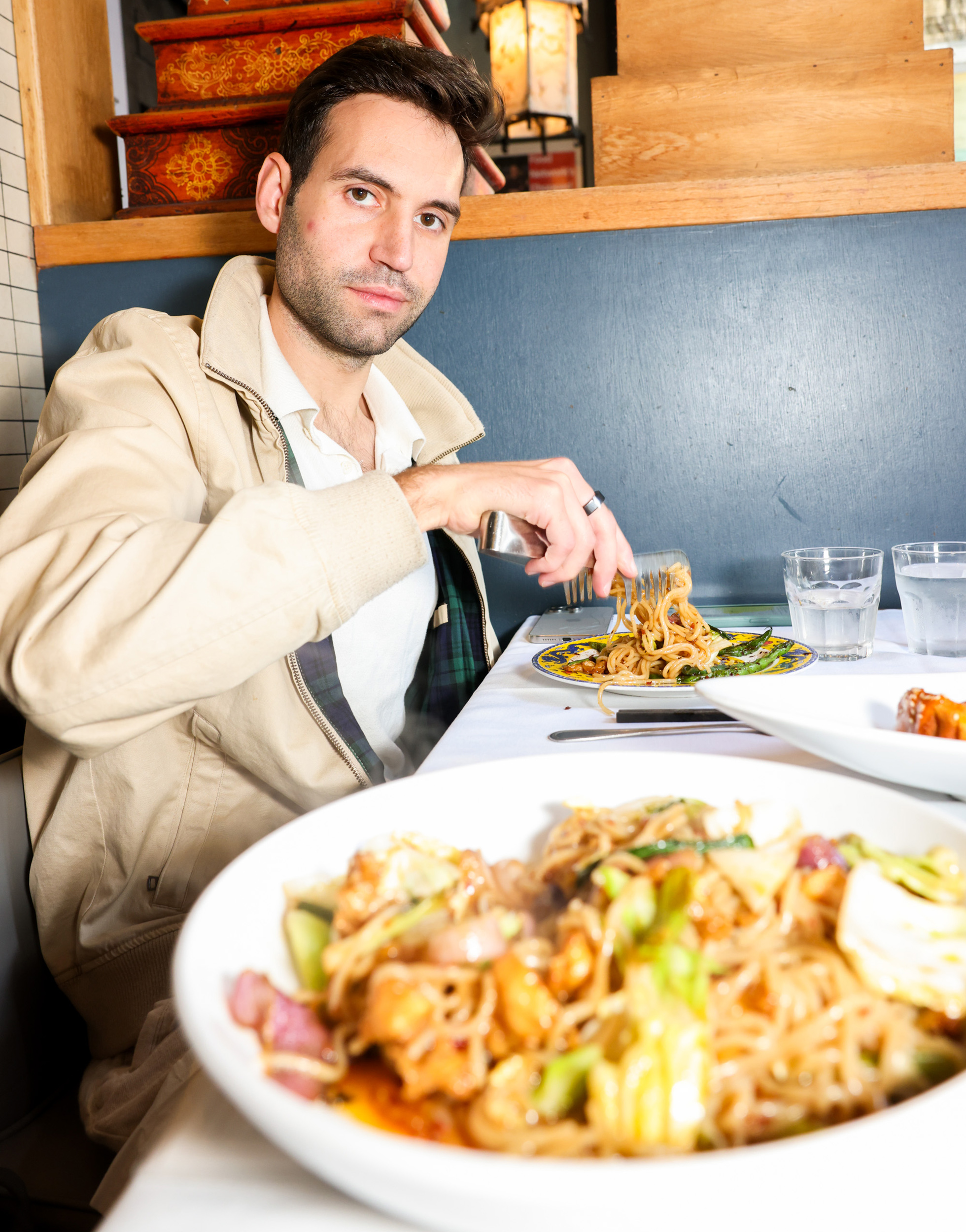 A person in a light jacket is seated at a table with a large bowl of noodles in front. They are holding a fork and looking at the camera, with more food and glasses of water visible.