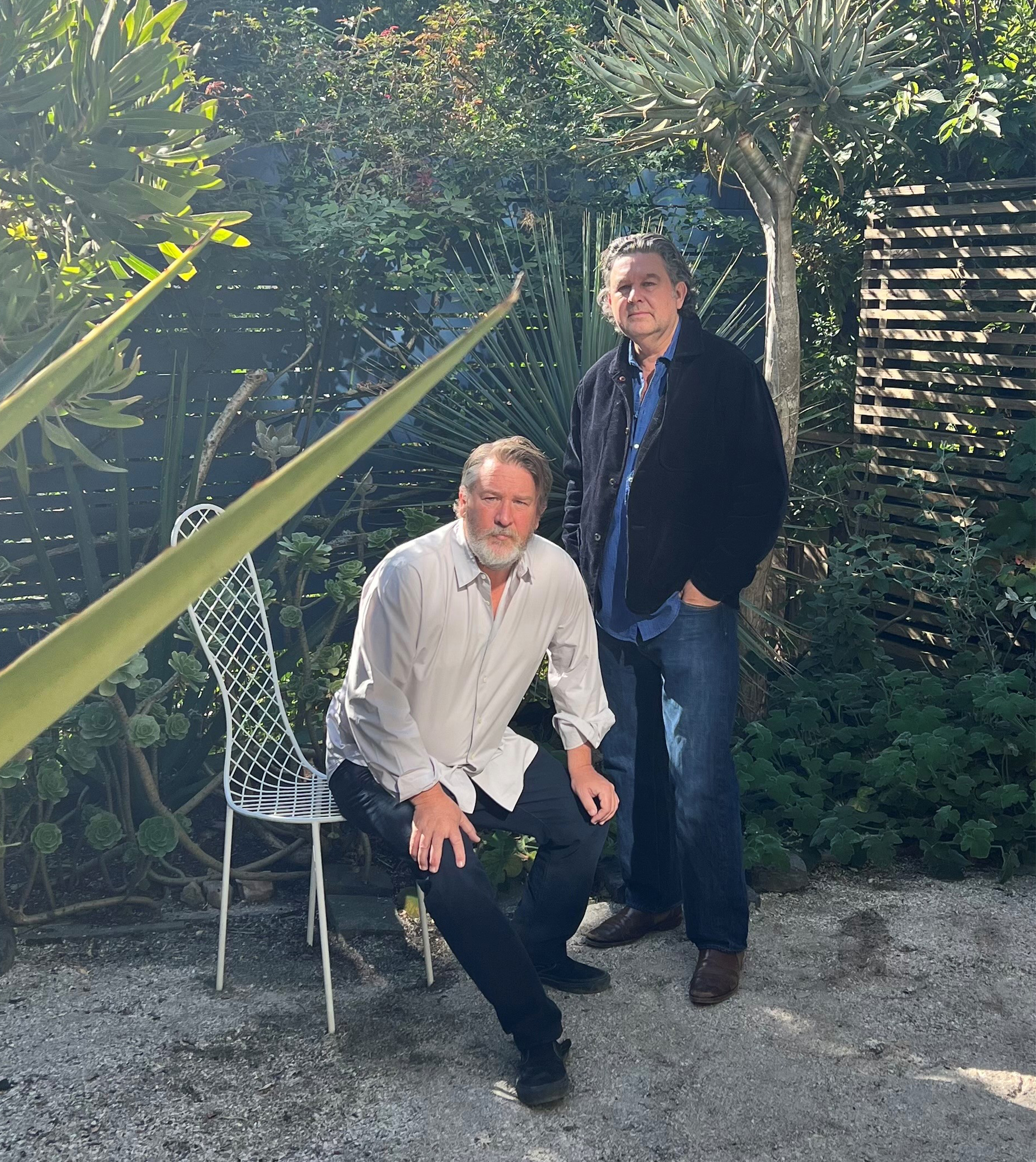 Two men are in a lush garden. One is sitting on a white chair, wearing a light shirt, and the other is standing, wearing a dark jacket and jeans.