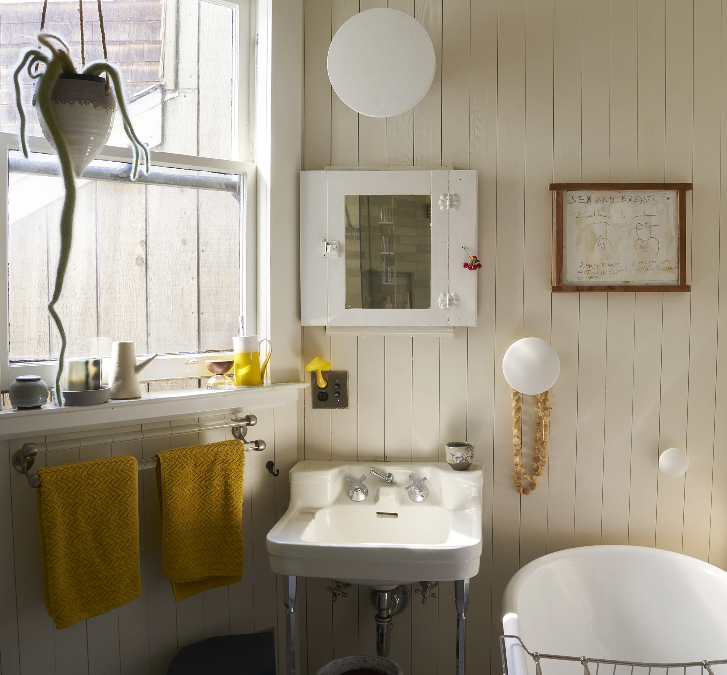 A bathroom with white walls features a sink with silver faucets, yellow towels on a rack, a bathtub, a hanging plant, and decorative wall items.