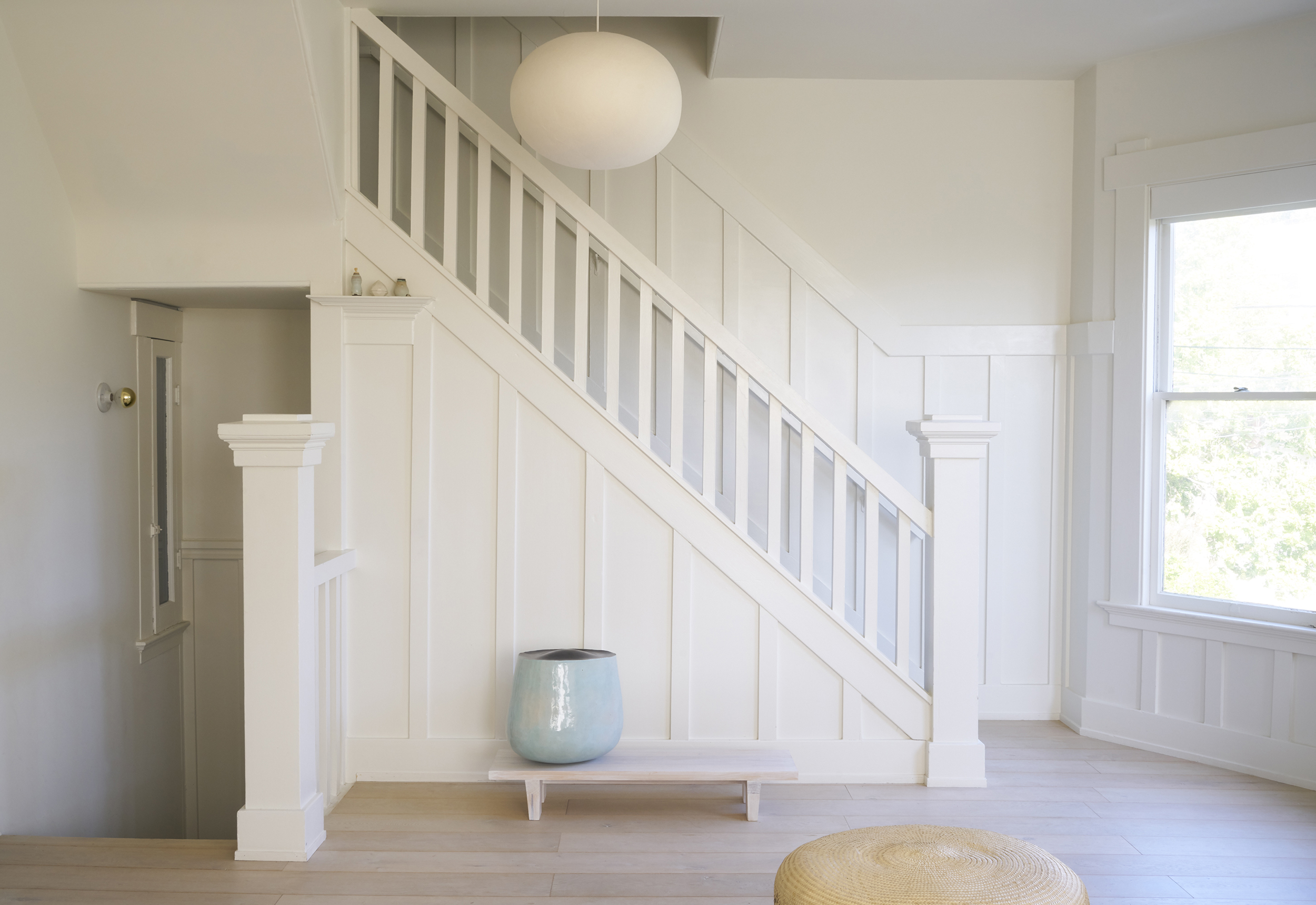 A minimalist stairway with white walls and railing, a round pendant light, a large window, a blue vase on a small bench, and a woven ottoman on light wood floors.