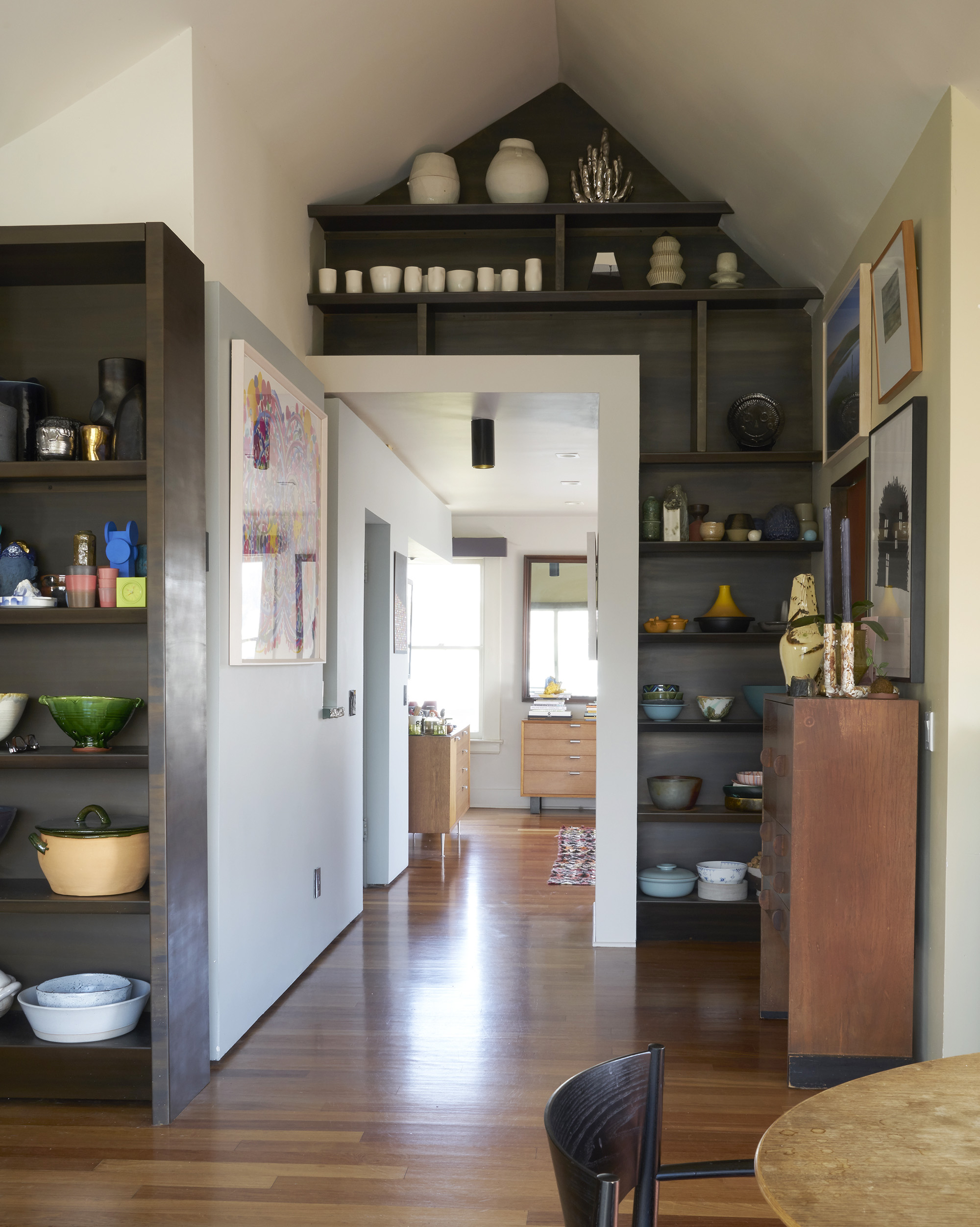 A cozy hallway with wooden floors, built-in shelves filled with pottery, and artwork on walls. The room is well-lit with natural light from a distant window.