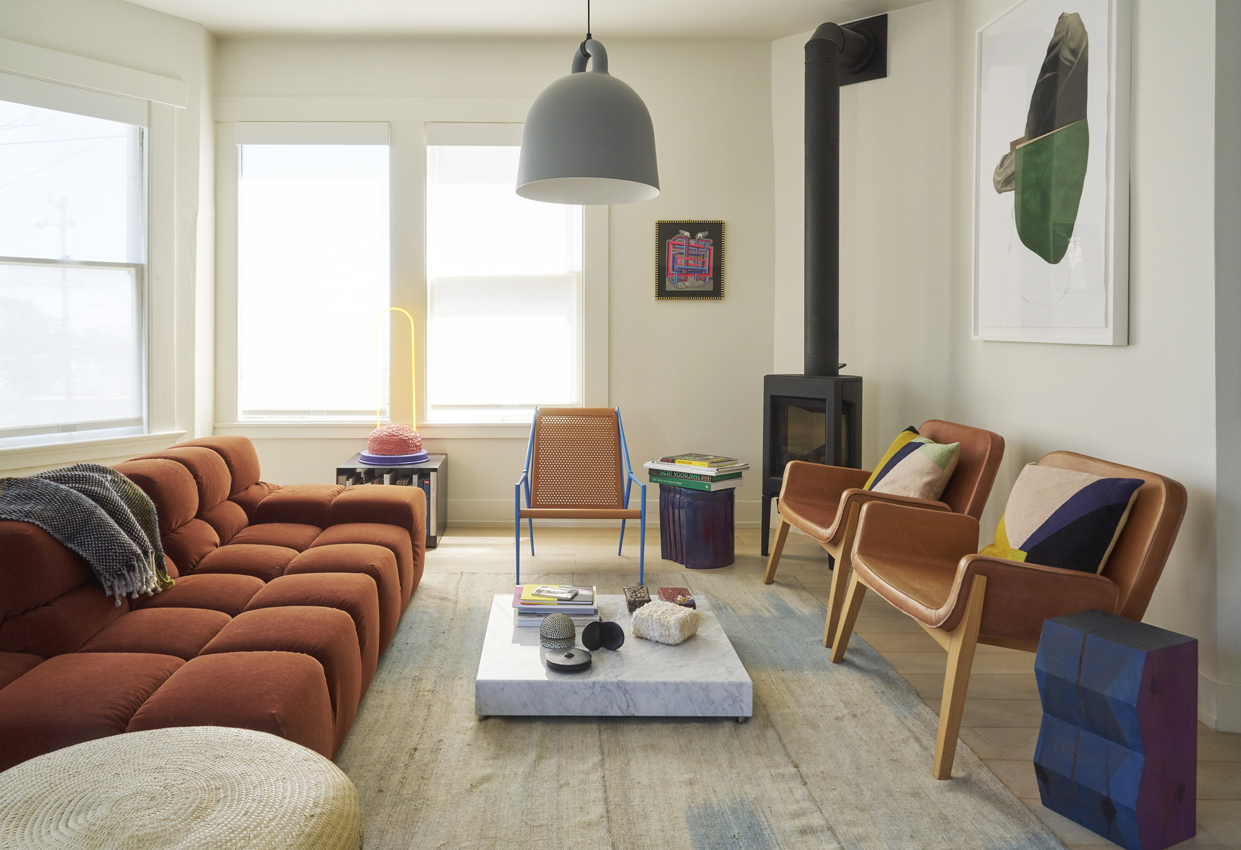 A modern living room with a rust-colored sofa, two tan chairs, a marble coffee table, and a black wood stove. Bright artwork and decor add color.