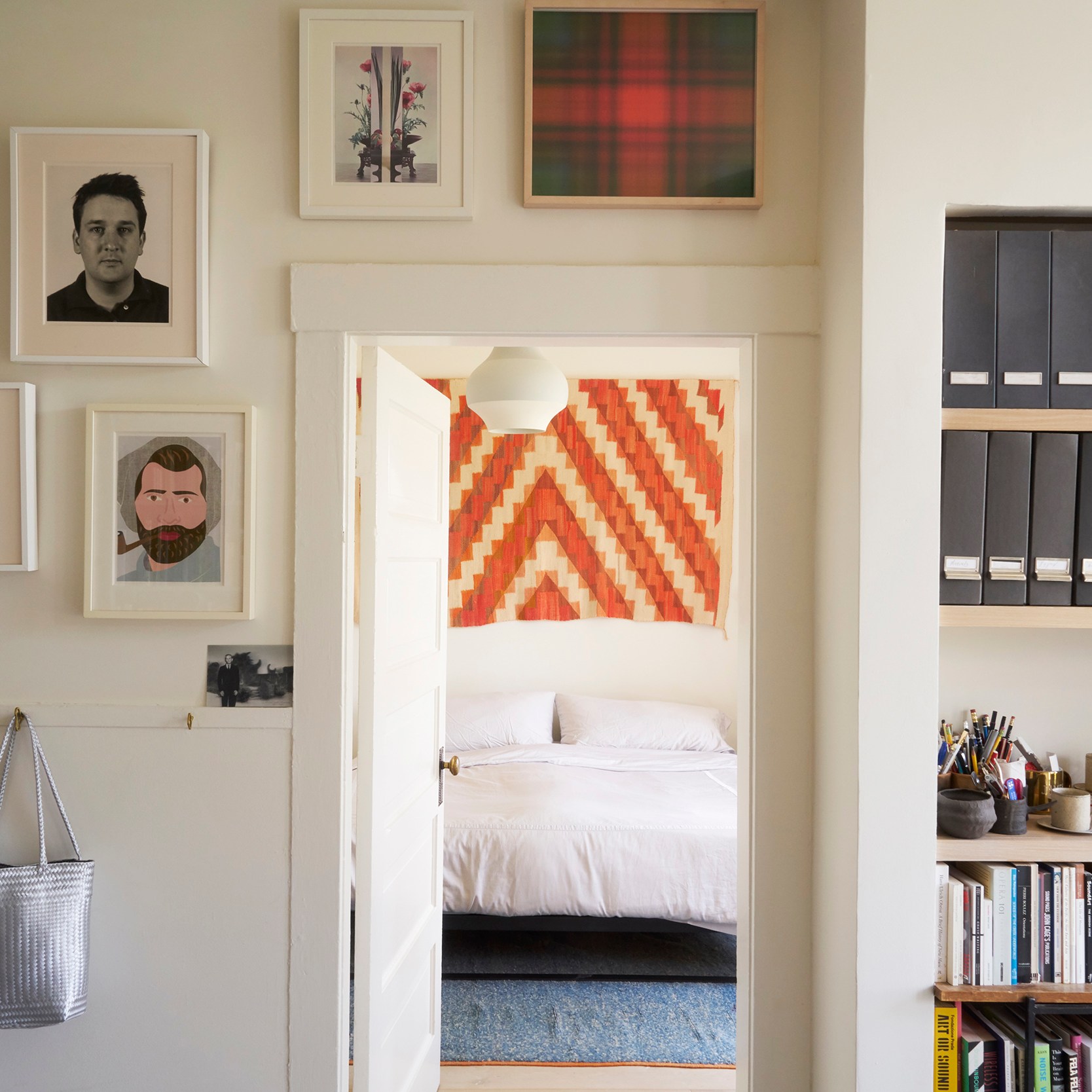 A cozy room with art on the walls, a door leading to a bedroom with a patterned decoration, shelves with files and books, and bags hanging on the walls.