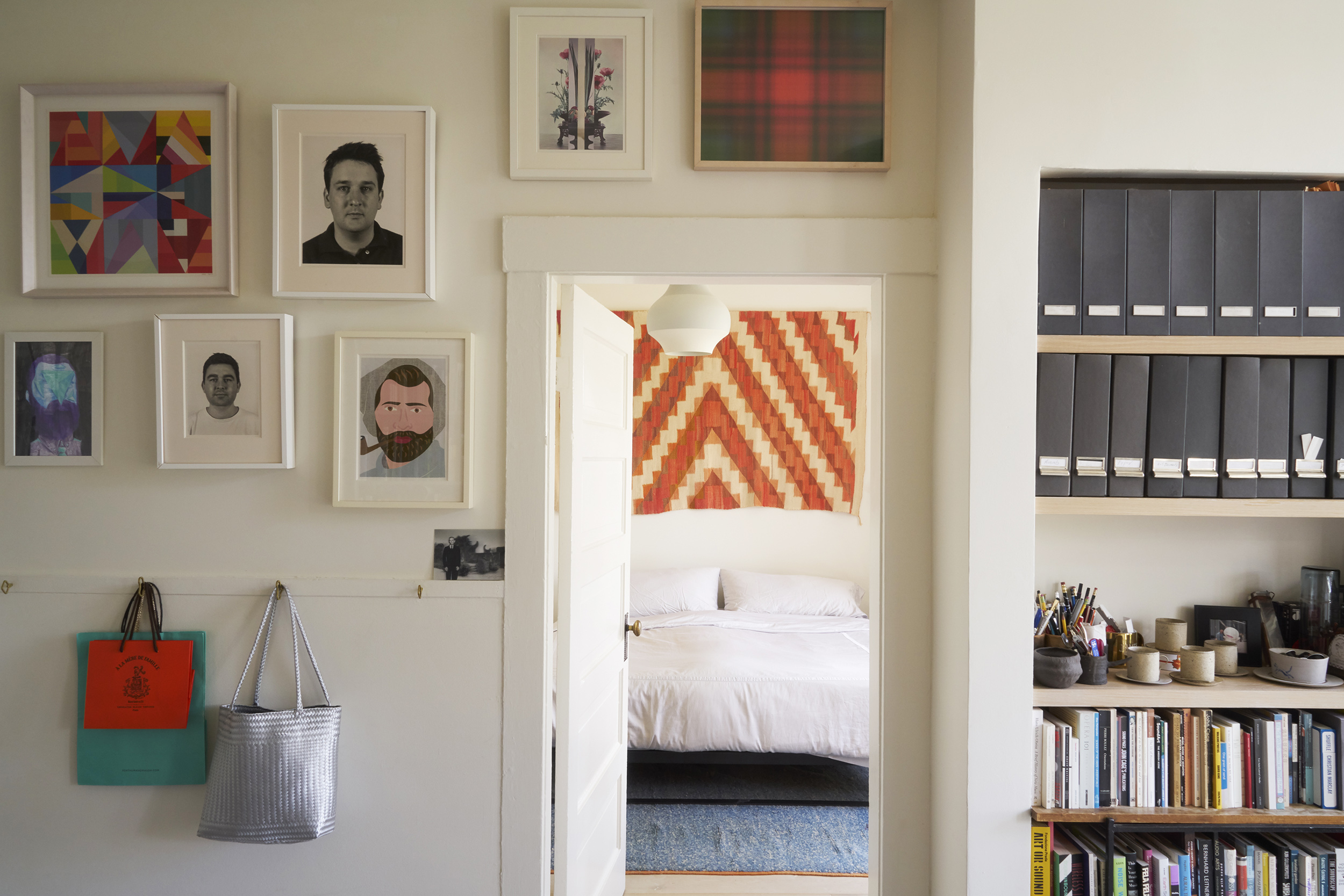 A cozy room with art on the walls, a door leading to a bedroom with a patterned decoration, shelves with files and books, and bags hanging on the walls.