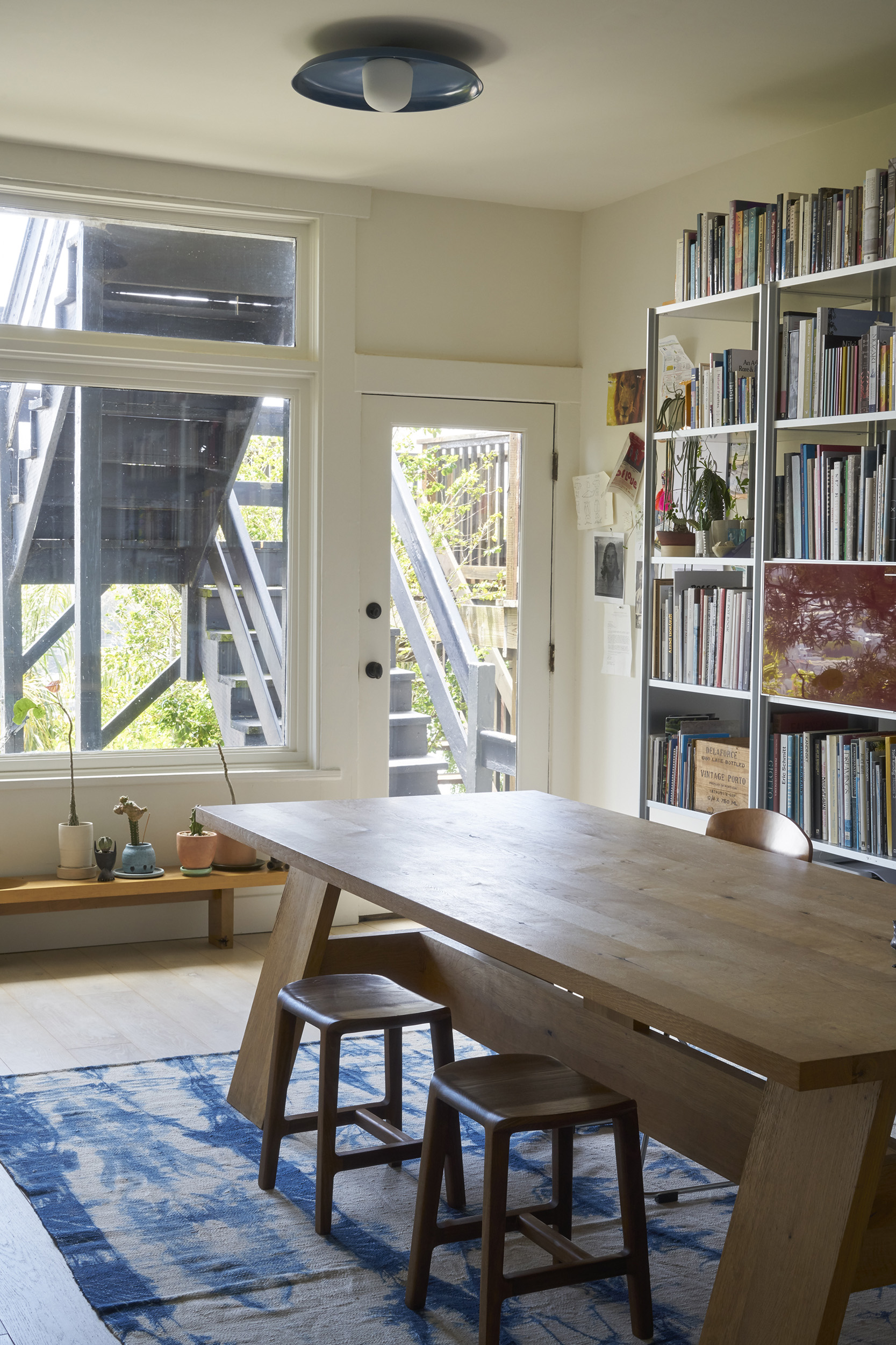 The image shows a cozy study with a wooden table, stools, bookshelves, and a door leading outside. Natural light streams in, enhancing the serene atmosphere.