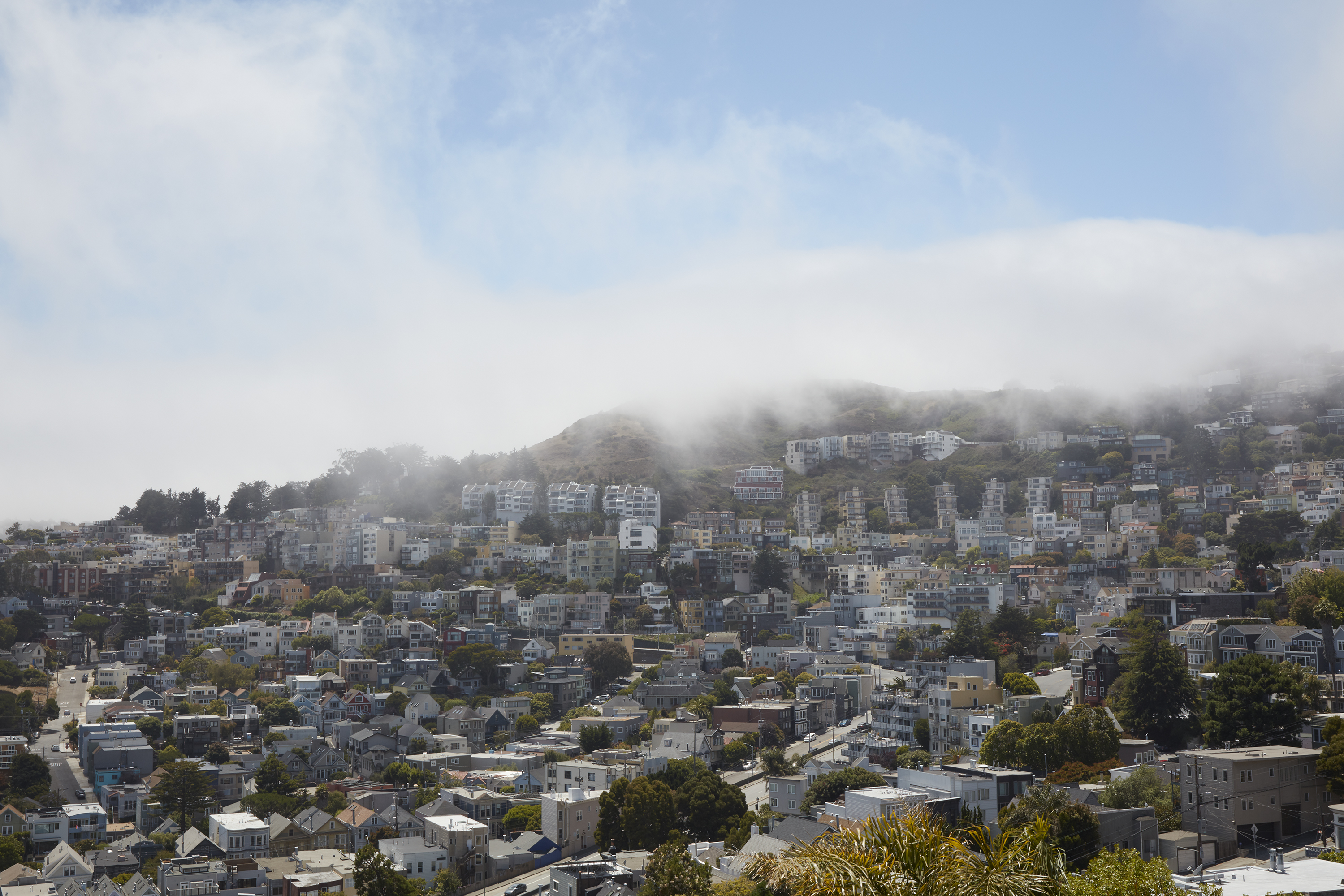 A hilly urban landscape with numerous houses and buildings. Fog partially covers the hills, and the sky is mostly clear with a few clouds.