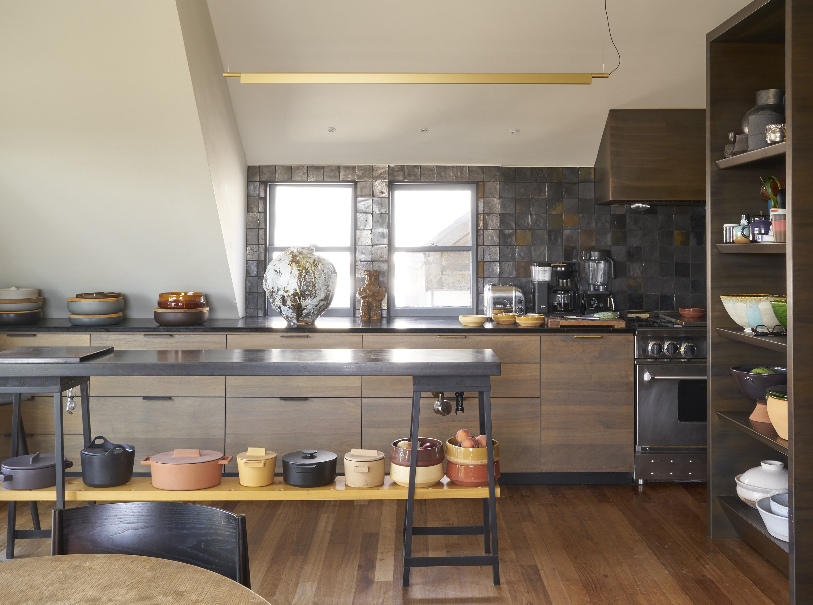 A modern kitchen features wooden cabinets, a dark countertop, pottery, a coffee machine, and a toaster. Shelves hold colorful bowls, and two large windows add light.