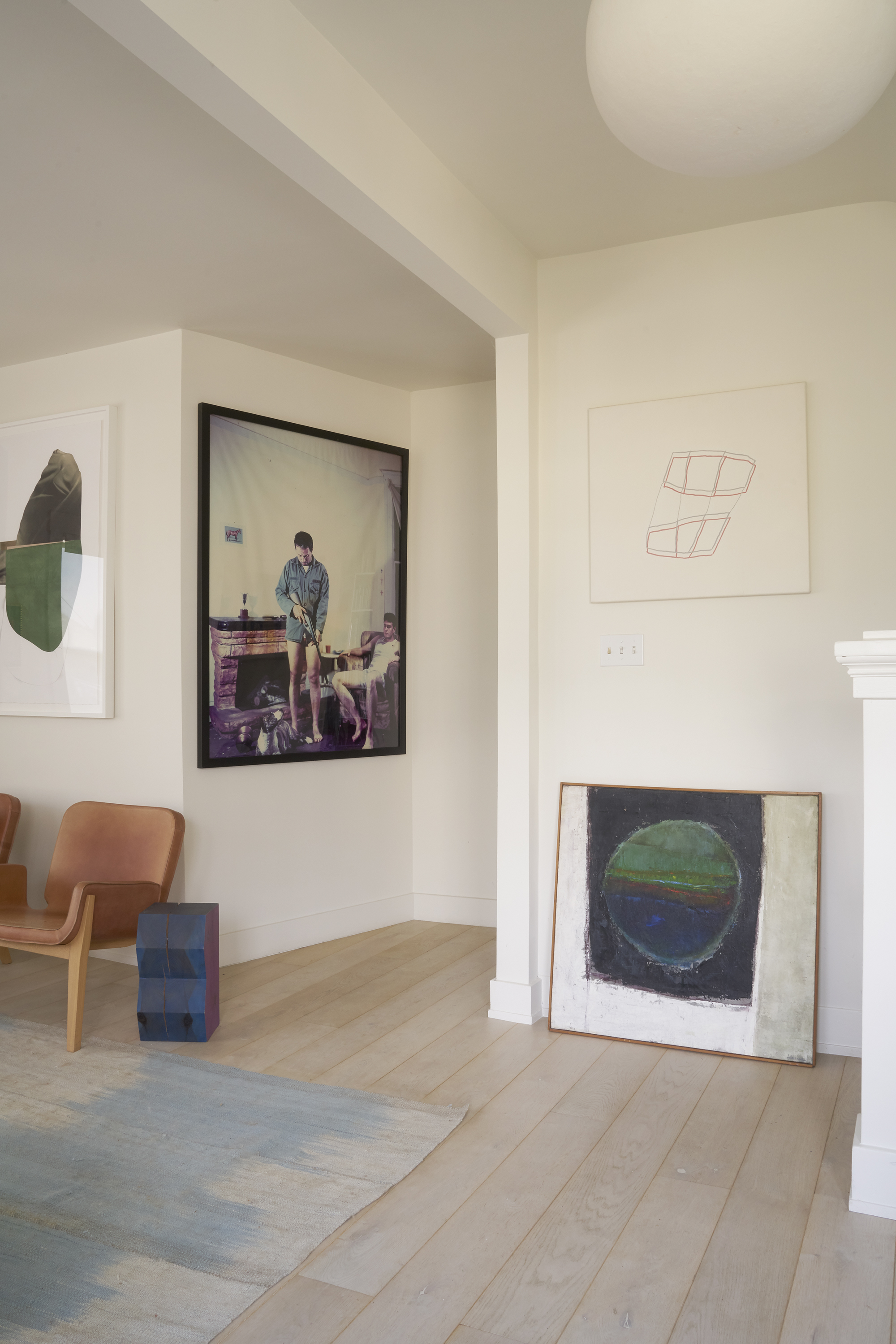 A modern interior with light wood flooring, an abstract rug, a brown chair, and eclectic artworks on walls, including a large framed photo and abstract prints.