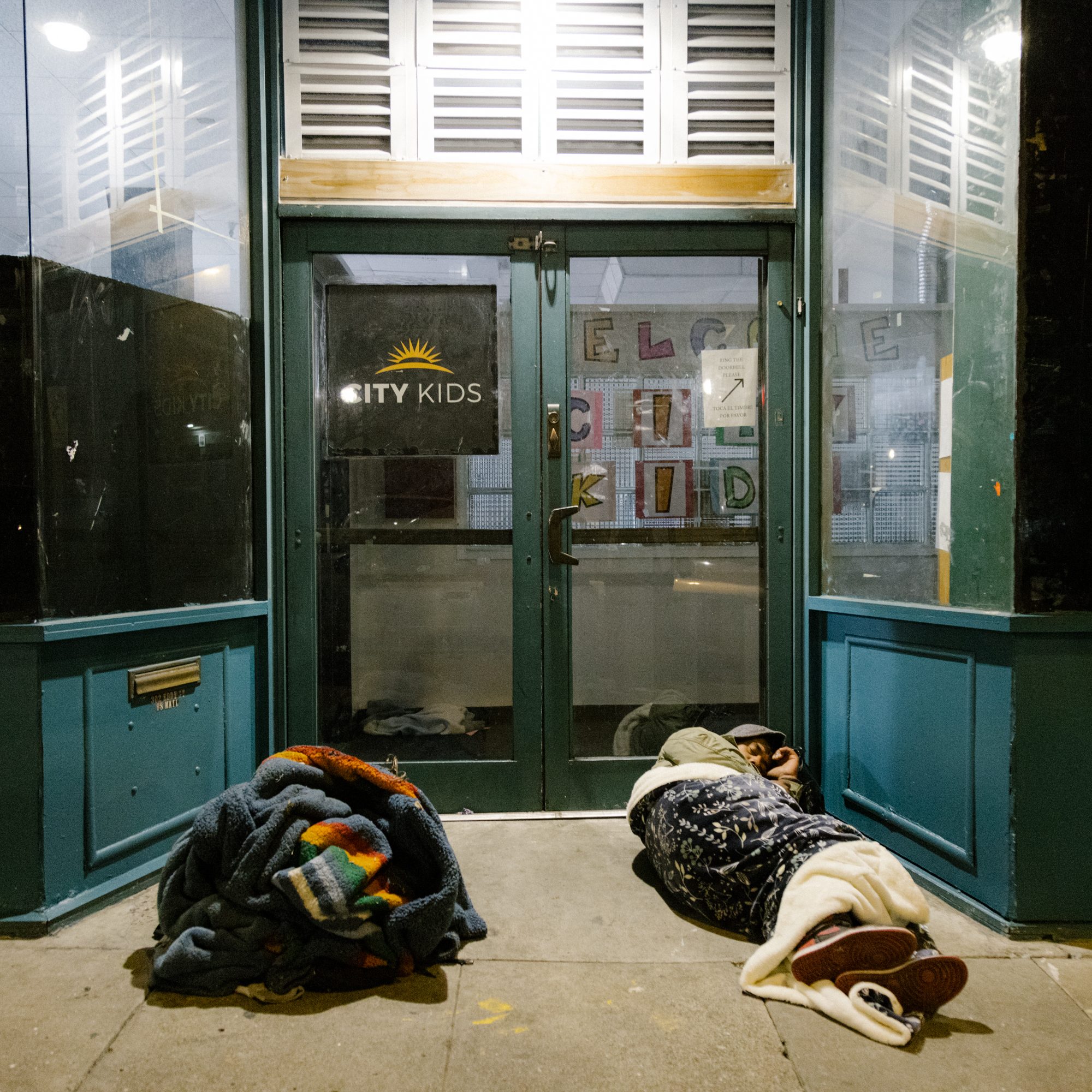 Two people are sleeping on a sidewalk, wrapped in blankets, in front of a storefront with a &quot;City Kids&quot; sign and colorful posters inside.