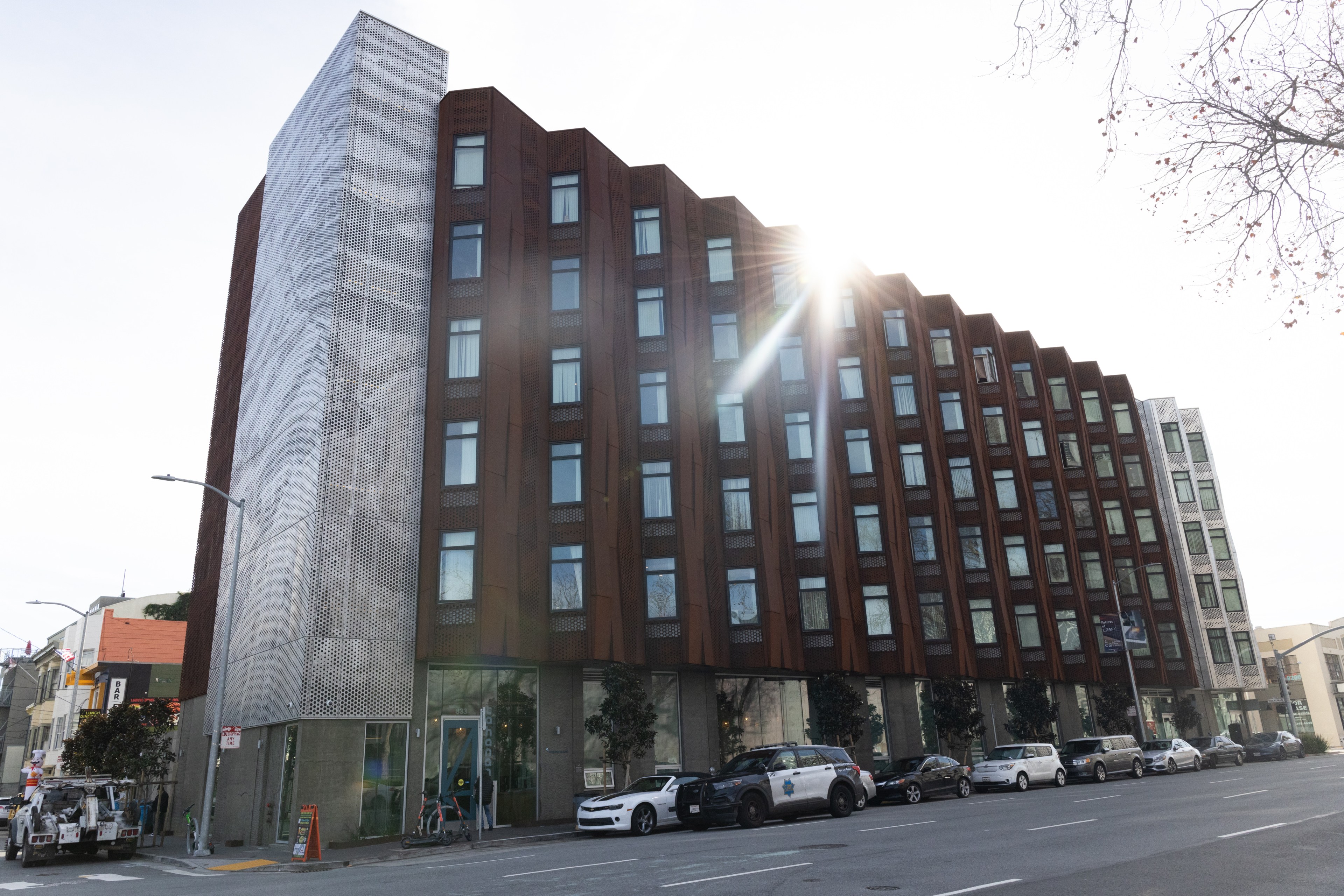 A modern building with a metal facade and large windows is on a city street. The sun shines above, and several cars are parked in front.