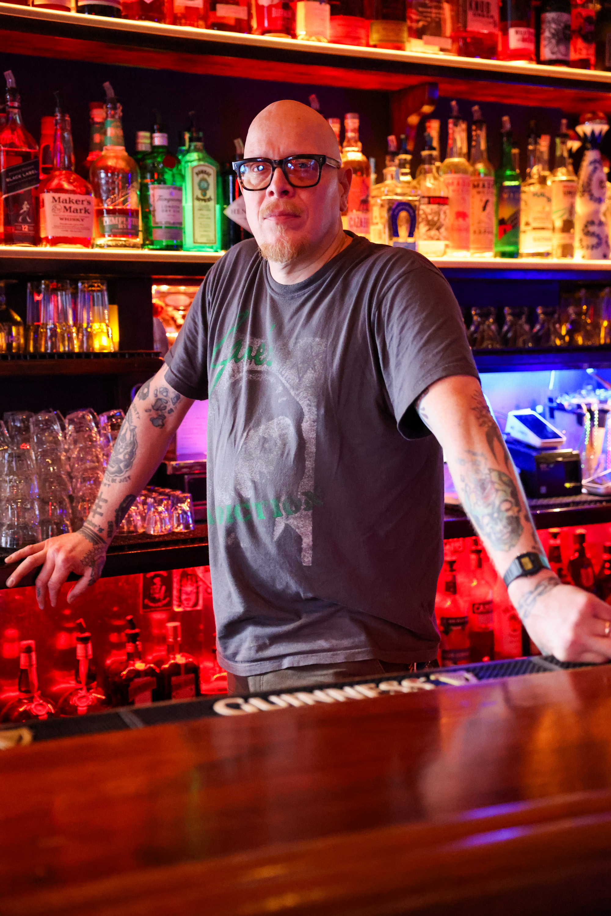 A bald man with tattoos, wearing glasses and a gray t-shirt, leans on a bar counter surrounded by colorful bottles and glowing lights.