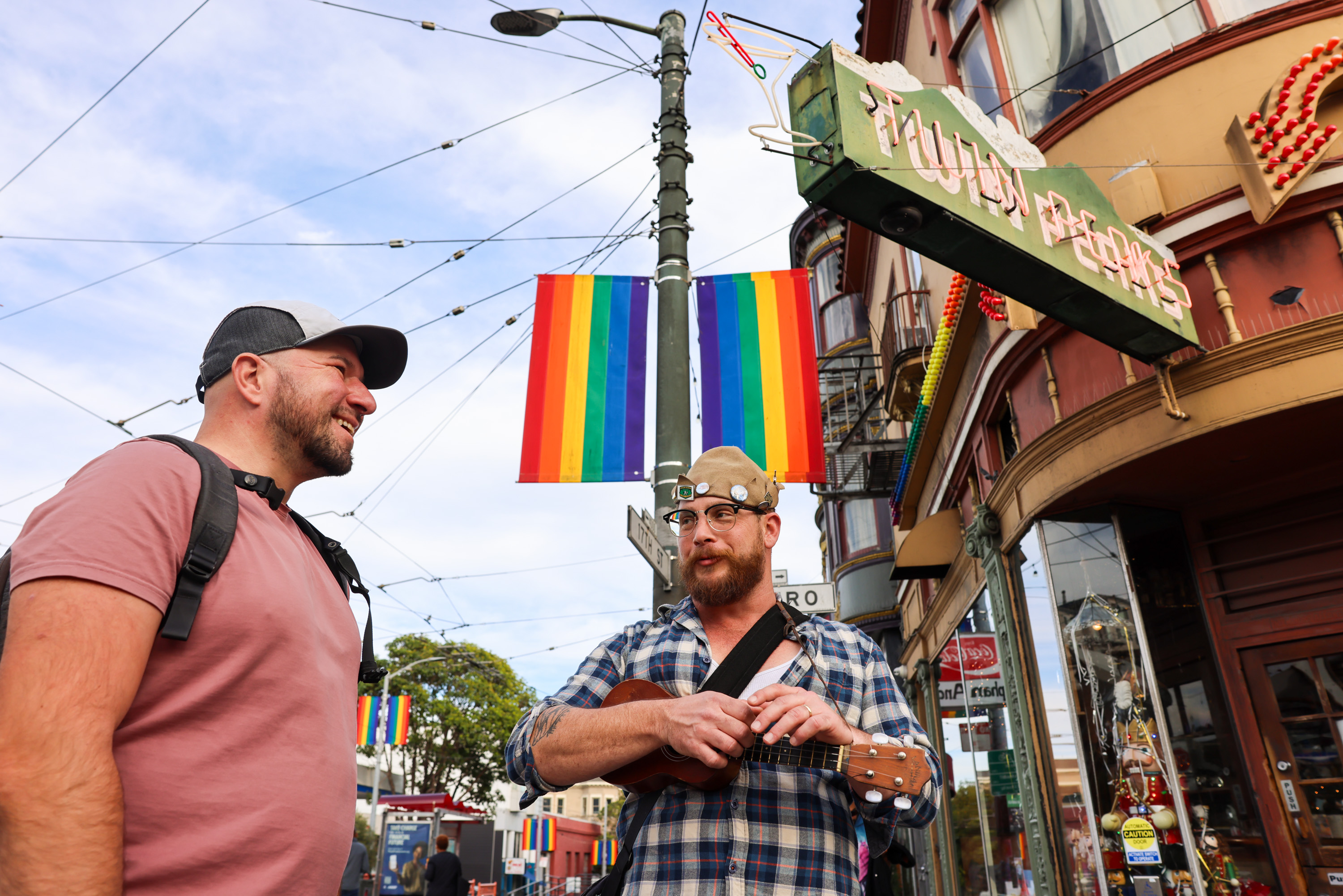 Two men stand outside a colorful building, one playing a ukulele. A rainbow flag hangs from a street pole, and string lights crisscross above their heads.