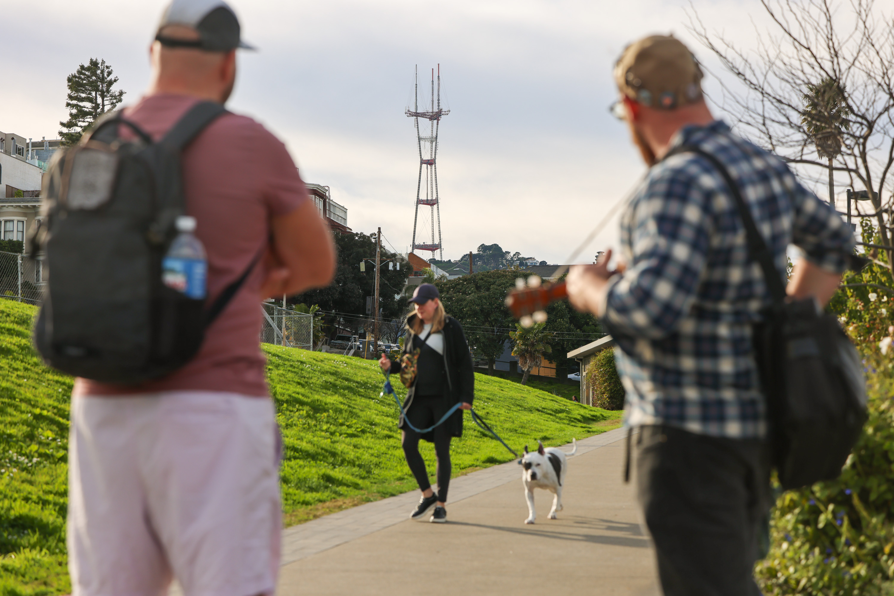 A woman walks her dog on a path with a tower in the background. Two men are in the foreground, one with a backpack and the other playing a guitar.