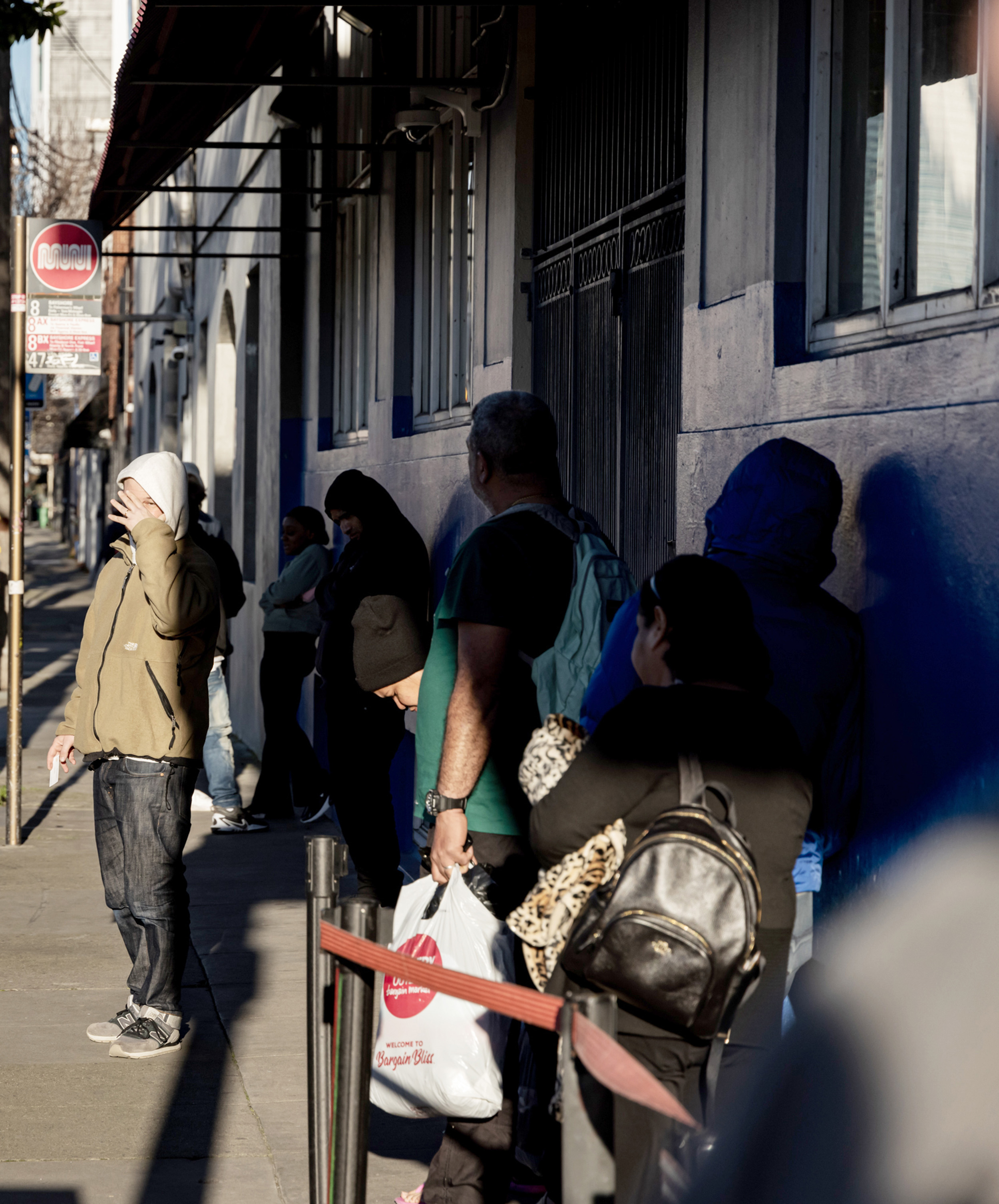 People are standing in line outside a building on a sidewalk. Some are dressed warmly, with hoods and jackets. One person holds a shopping bag.