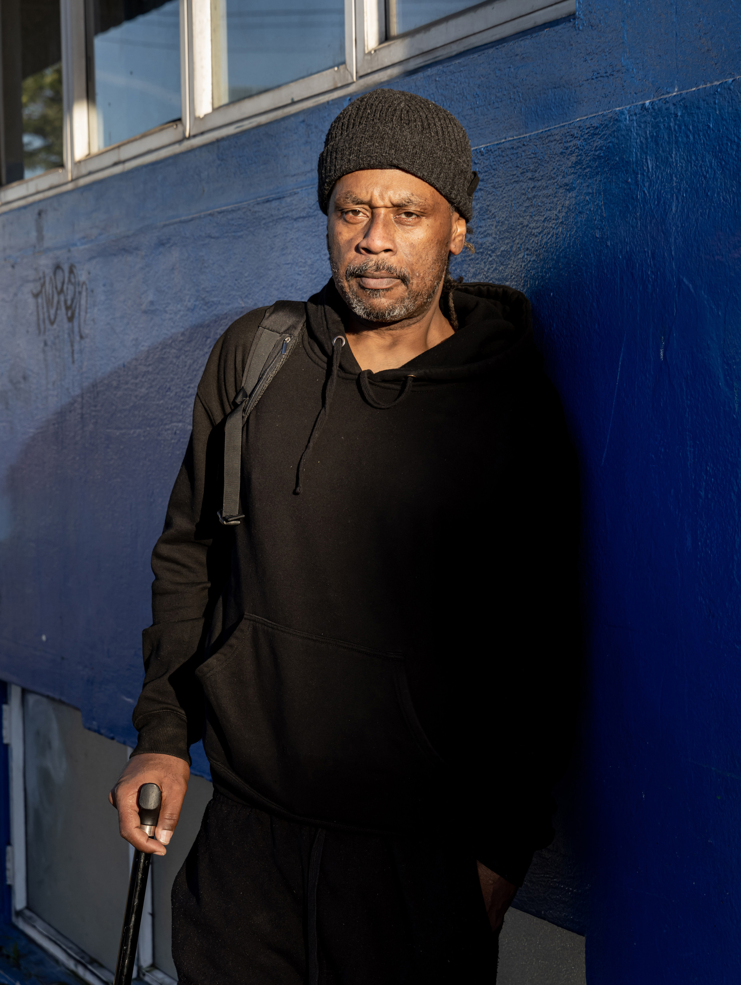 A man wearing a black hoodie and beanie stands against a blue wall, holding a cane. Sunlight highlights his face as he looks directly ahead.