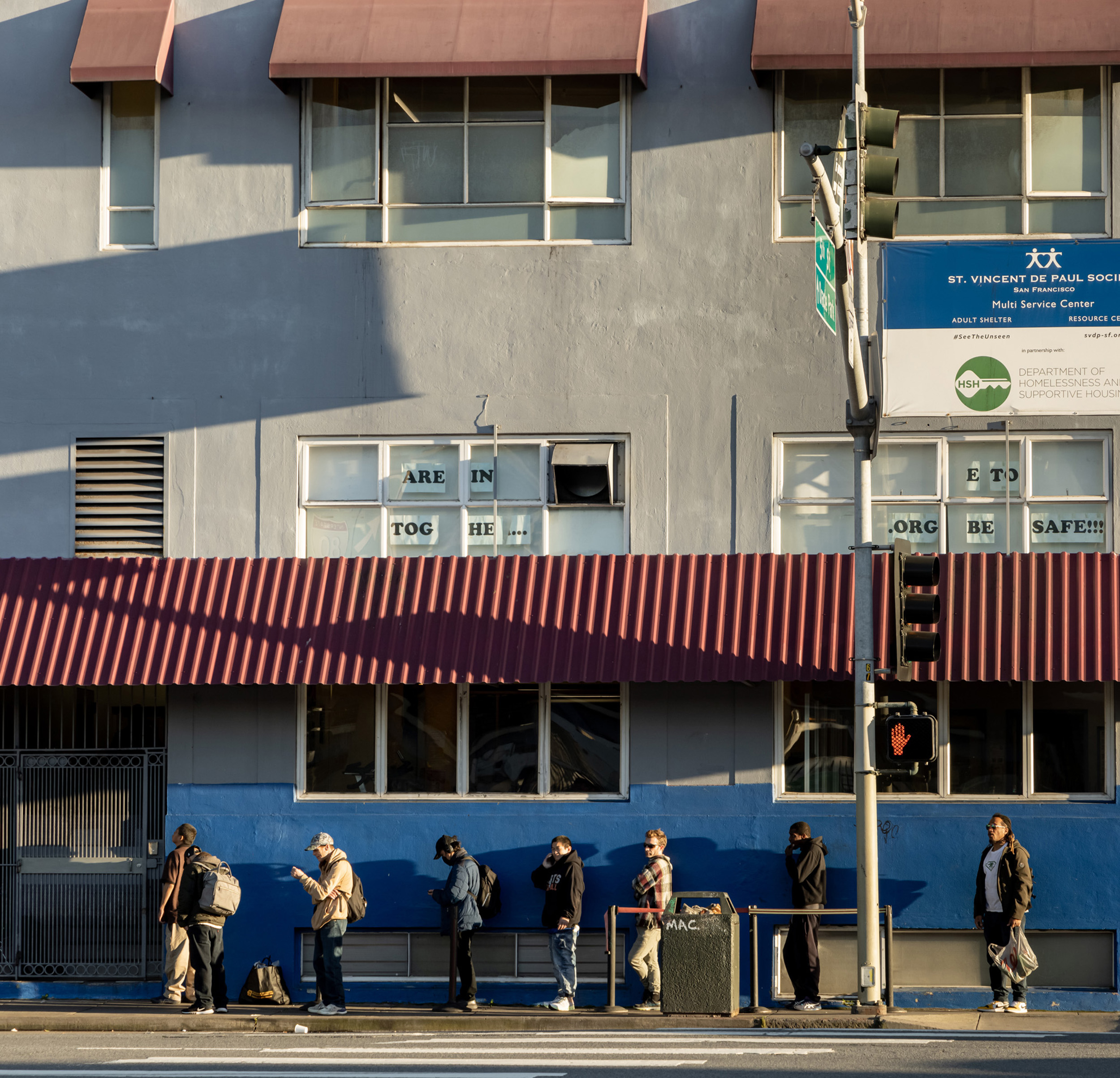 People stand in line outside a building labeled &quot;St. Vincent de Paul Society&quot; with signs reading &quot;ARE IN TOG HE...&quot; and &quot;BE SAFE!!!&quot; under a red awning.