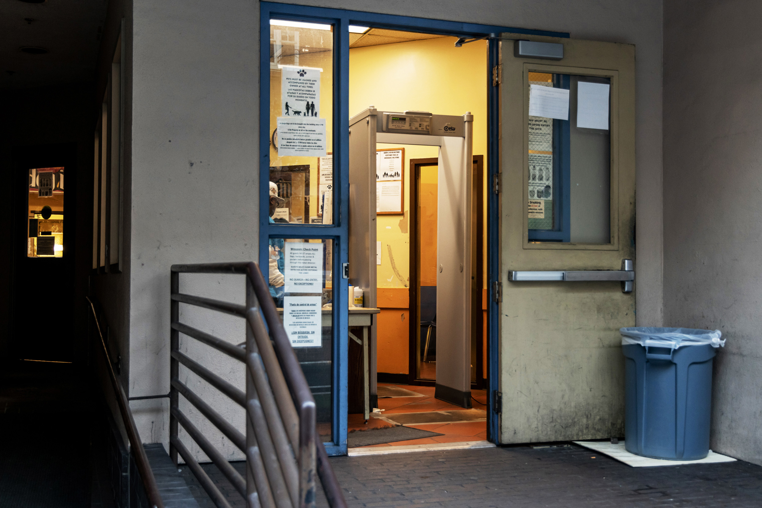 The image shows an open doorway leading to a room with a metal detector. Notices are posted on the glass, and a trash can is placed outside.